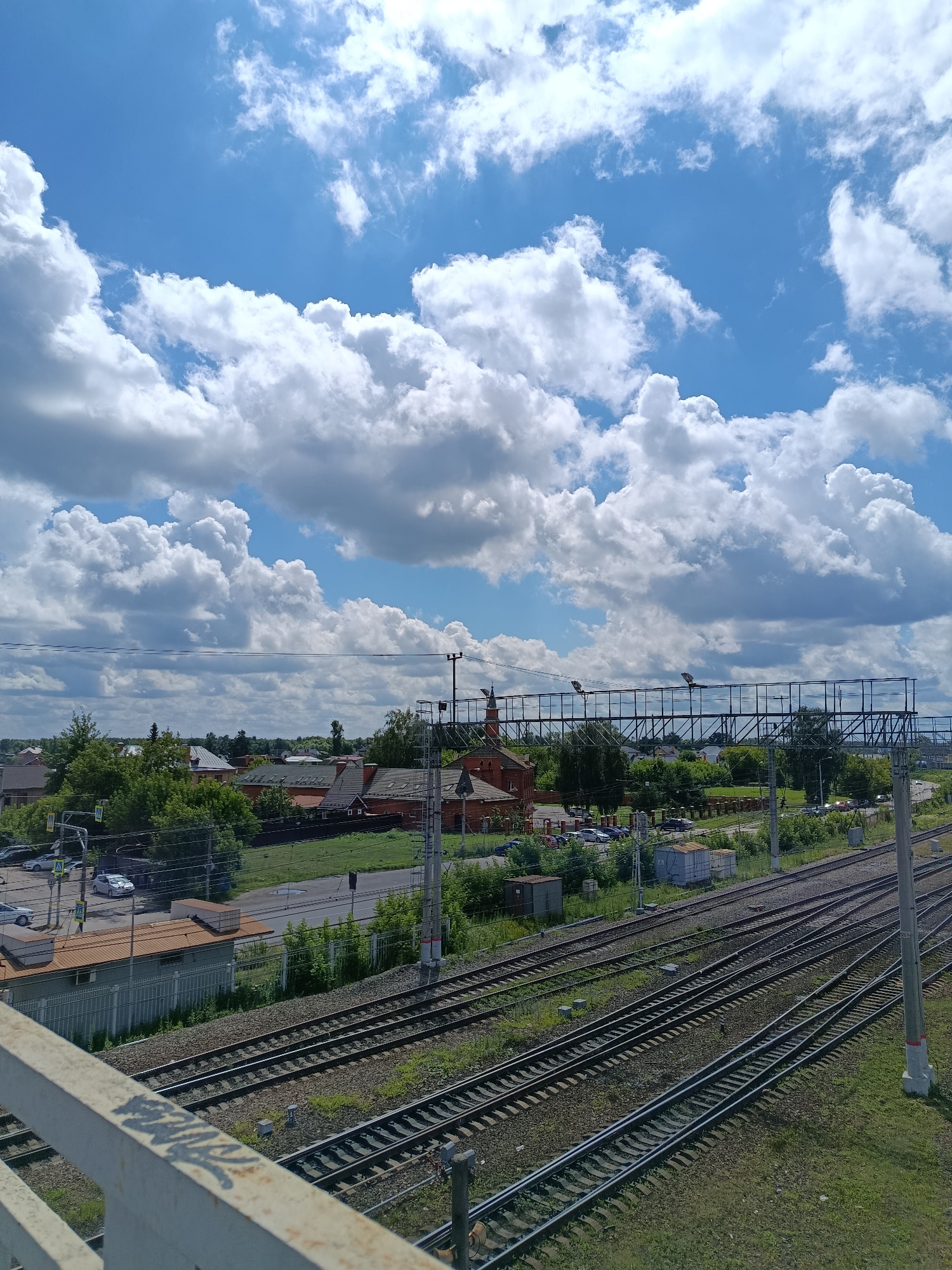 This is an eye-level, medium-shot photograph taken from an elevated position, possibly a pedestrian overpass, looking across multiple railroad tracks. The scene is set in the city of Orekhovo-Zuyevo, Russia.
In the foreground, a white metal railing, tagged with graffiti, is visible on the bottom left. Beyond the railing, several parallel railroad tracks stretch diagonally from the bottom right towards the center of the image and recede into the distance. The tracks are surrounded by green grass and gravel.
In the midground, a town or village is situated to the left of the tracks. Several houses with red-tiled roofs and brick buildings are visible, interspersed with lush green trees. A red brick building, possibly a church or a public building with a prominent steeple, stands out among the structures. Adjacent to the houses is a road with several parked cars. Overhead, a network of power lines and structures supporting them are present.
The background of the image is dominated by a bright blue sky filled with large, fluffy white cumulus clouds. The clouds are dense and voluminous, creating a dramatic and dynamic sky. The overall lighting suggests a sunny day. The scene depicts a typical urban or suburban landscape in Russia, with a focus on the infrastructure of the railway system and the surrounding residential area under a vast, cloud-filled sky.