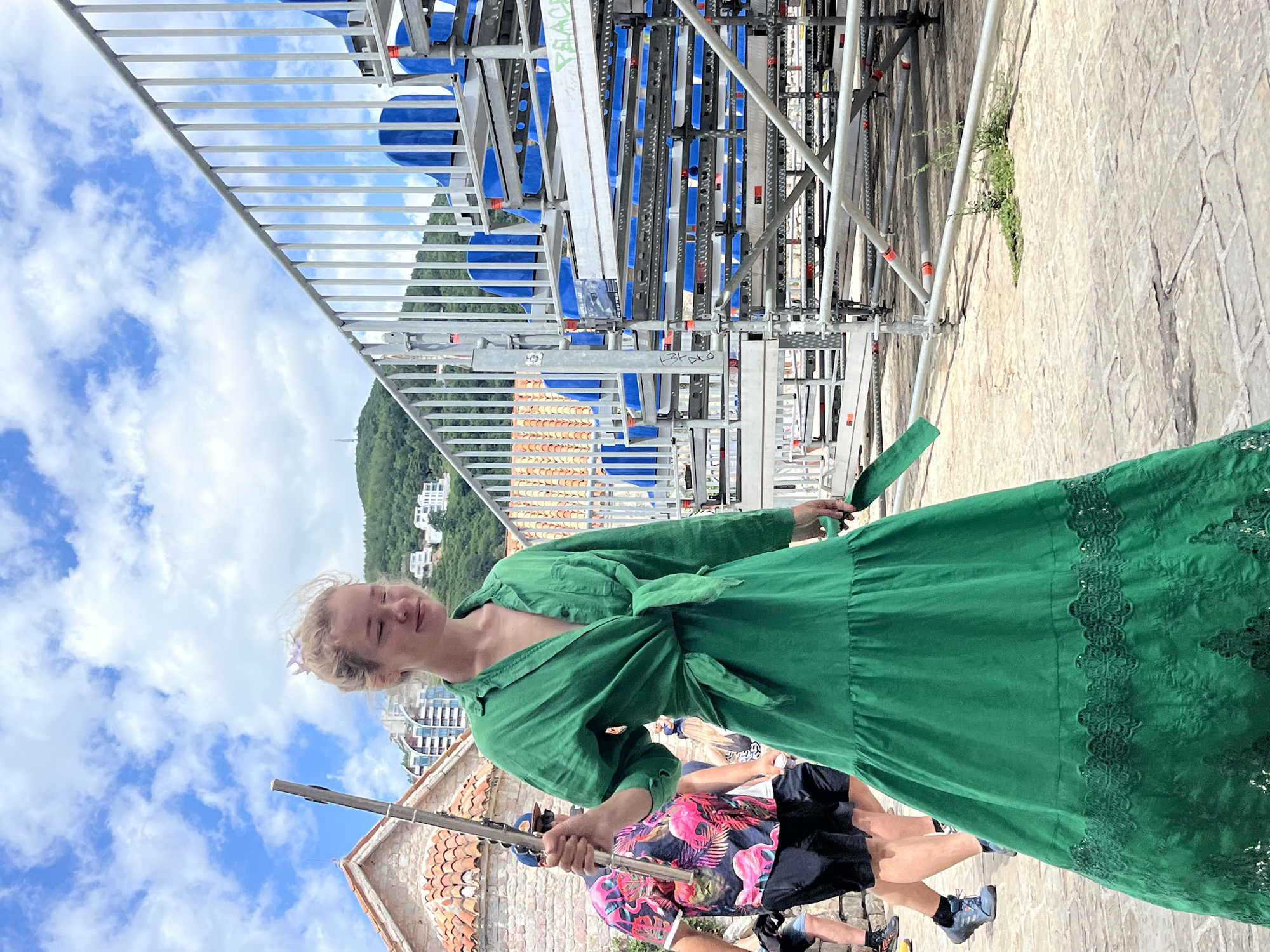 The image depicts a woman in a green dress standing on a stone pathway, possibly outdoors. The pathway is flanked by scaffolding structures, with blue elements visible within the scaffolding. In the background, there is a lush green hillside dotted with buildings, and a bright blue sky with scattered clouds. The woman is looking down and to her left, with a slight smile on her face. She has blonde hair, and a small purple flower is tucked behind her ear. Her dress is long, with lace detailing on the hem. She is holding a wooden pole in her right hand. There are other people visible in the background, one of whom is wearing a shirt with a flamingo pattern. The overall impression is of a warm, sunny day, possibly in a Mediterranean location given the architectural style of the buildings and the lush vegetation. The scaffolding suggests some ongoing construction or event setup.