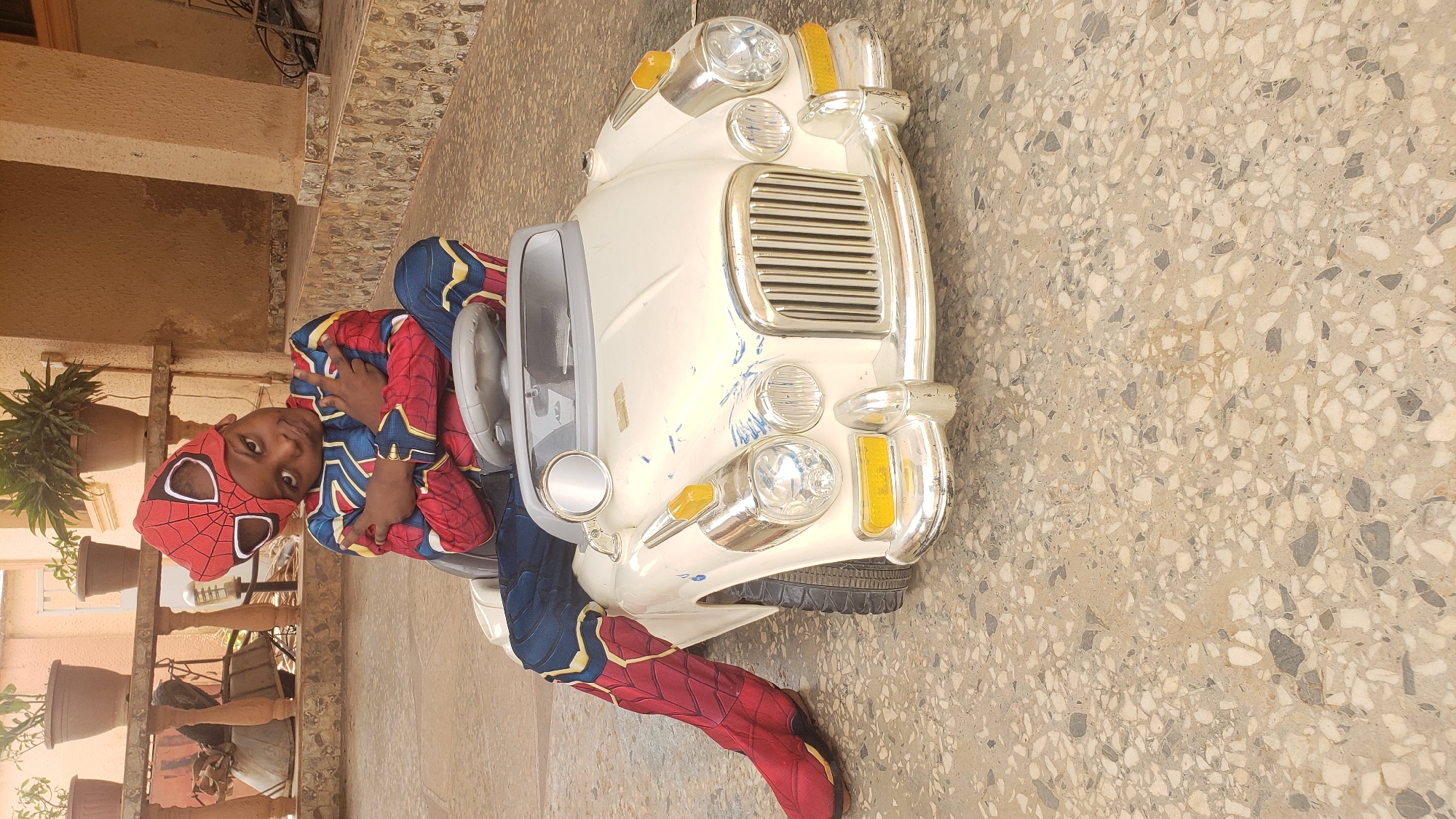 A young person dressed in a Spider-Man costume is sitting in a white toy car outdoors. The ground is a beige terrazzo surface. The background shows a building with plants in pots on a balcony, located somewhere in Sabon Tasha, Nigeria.