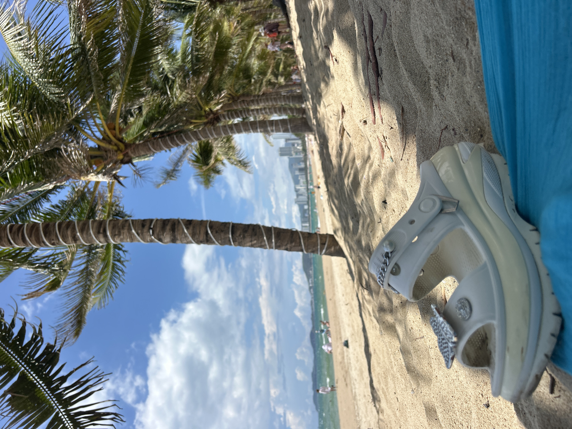 The photograph shows a beach scene in Nha Trang Ward, Vietnam. Palm trees line the beach, with a view of the ocean and buildings in the distance under a blue sky with clouds. A white Croc sandal with decorative charms lies on the sandy beach near a blue object. People are visible in the background along the shoreline.