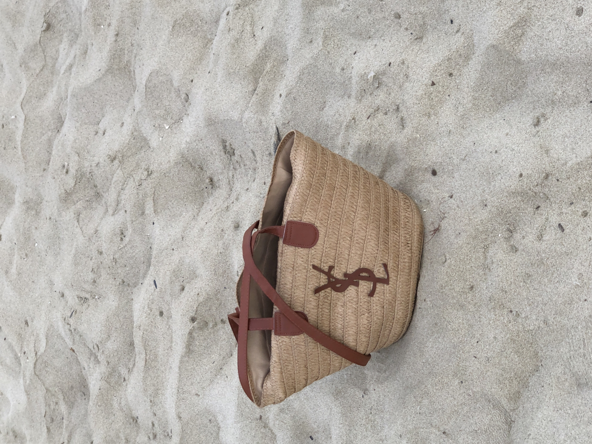 A straw tote bag with brown leather trim and handles sits on a sandy surface. The bag has the Yves Saint Laurent logo on the front. The sandy surface appears to be a beach, with some small shells and debris scattered across it. The lighting suggests it is daytime. This scene could be at My Khe Beach in Đà Nẵng, Vietnam, a popular tourist destination known for its sandy shores.
