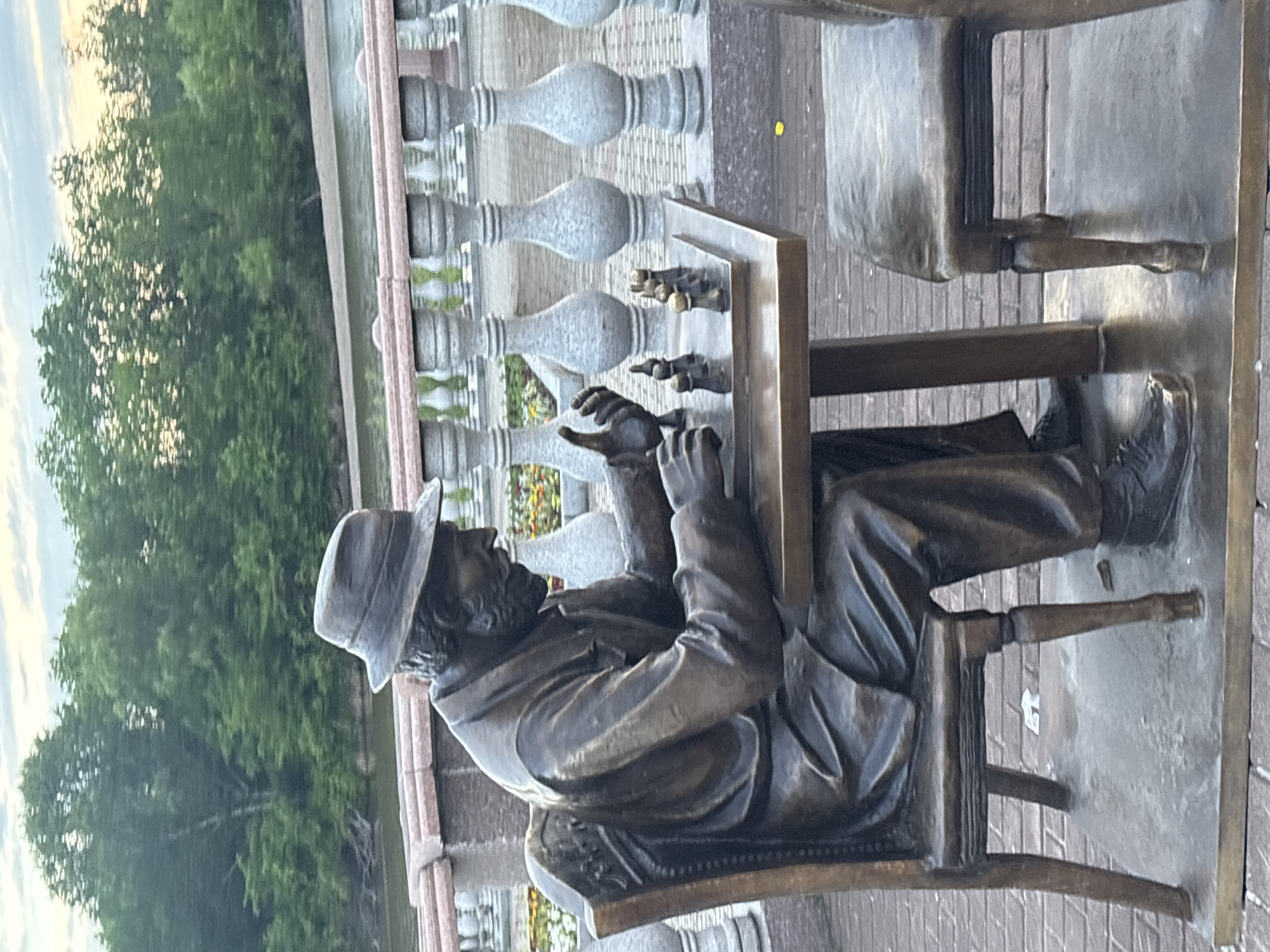 A bronze statue depicts a man in a hat and suit playing chess at a table. The statue is seated in a chair, facing a chessboard with pieces. Another empty chair is positioned across the table. The sculpture is situated outdoors on a brick pathway, with a decorative railing and trees visible in the background. The environment suggests a park or public space by a body of water.