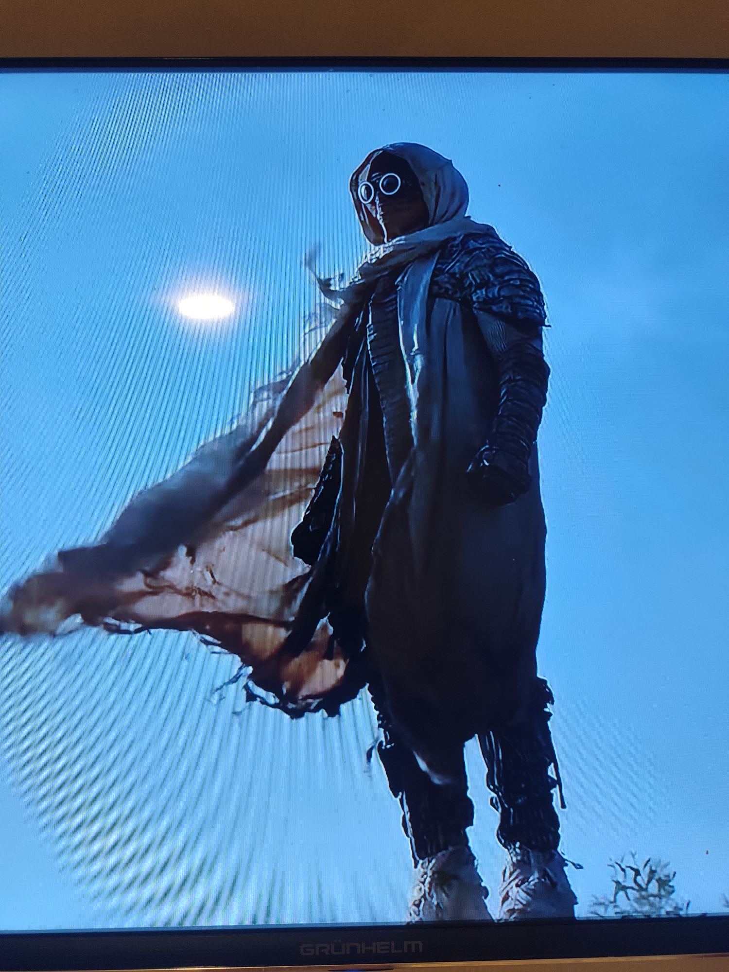 The image shows a low-angle shot of a person dressed in a hooded outfit with goggles. The person is wearing a large, flowing cloak with frayed edges that billows to the left. The outfit appears to be made of rough, textured materials, with layered armor on the shoulders and arms. The person's legs are covered in what looks like tattered trousers adorned with fringe or hanging strips, and they are wearing sturdy boots. The goggles are circular with white rims.
The background is a clear, bright blue sky, suggesting daytime. A soft, circular light source is visible in the upper left portion of the sky, possibly a reflection or a stylistic element. The overall aesthetic of the person's attire suggests a post-apocalyptic, fantasy, or sci-fi setting, perhaps in a desert or wasteland environment given the flowing garments and rugged appearance. There are no specific cues about the weather beyond the clear sky, and no discernible emotions are conveyed due to the obscured face.
At the bottom of the image, along the edge of the screen, the word "GRÜNHELM" is visible in white text against a dark background.