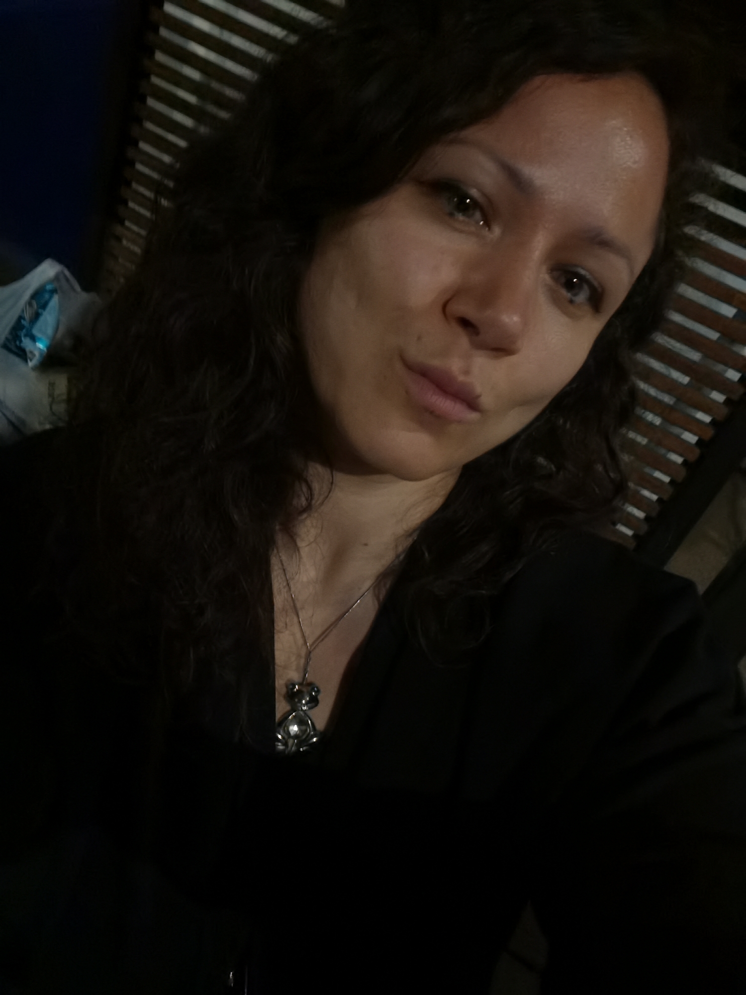 A woman with dark, wavy hair and light skin takes a selfie in a dimly lit room. She is wearing a black top and a silver necklace with a teddy bear pendant. The background features a slatted wooden structure and a blue bag. The photo appears to be taken indoors, possibly in a residential setting in Chaikovsky, Russia, given the presence of what might be a window blind made of wood. The lighting is low, creating shadows across the woman's face and the room.