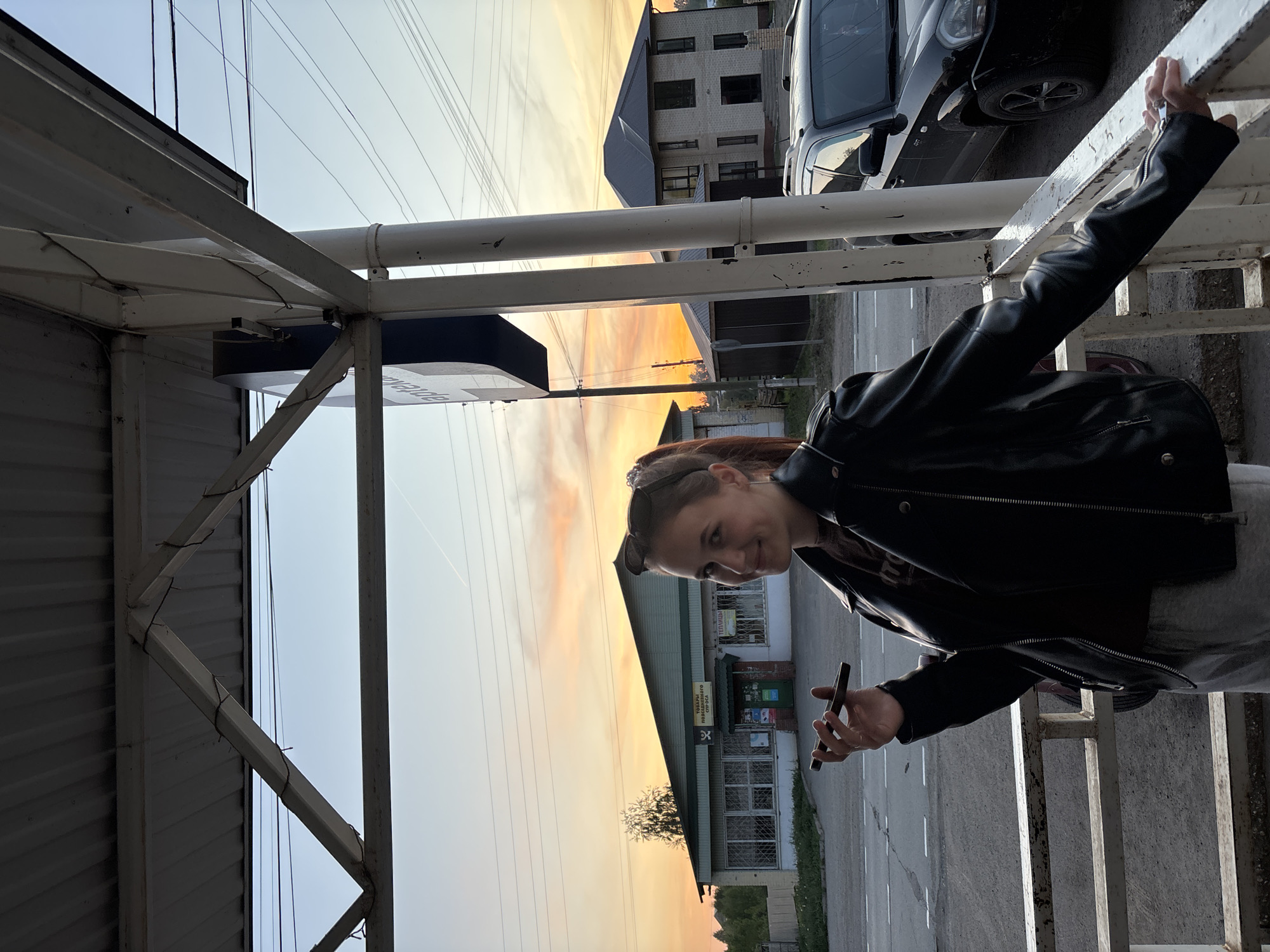 The image presents a woman in a black leather jacket, holding a smartphone. She stands on a metal railing, leaning against it with her right hand. Behind her, the setting sun casts an orange hue across the sky. Buildings are visible, including what appears to be a store, identifiable by Cyrillic script on its signage. A dark-colored car is parked nearby. The photo appears to be taken in Фоки, Russia.