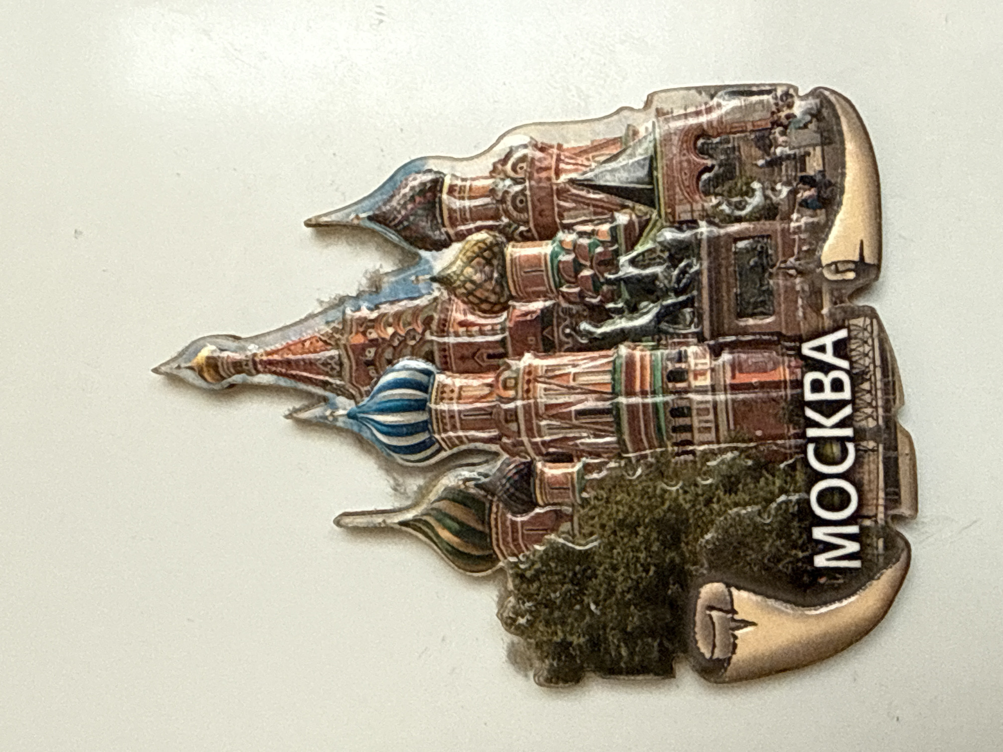 The image is a close-up, top-down shot of a magnet shaped like St. Basil's Cathedral in Moscow. The magnet is a colorful, detailed replica of the iconic landmark, featuring its distinctive onion domes and intricate architectural elements. The text "МОСКВА" (Moscow) is printed in white capital letters on the lower left portion of the magnet. The magnet is positioned on a plain white surface, with some subtle shadows cast by its edges, indicating a light source from above. The overall impression is that of a souvenir item.
