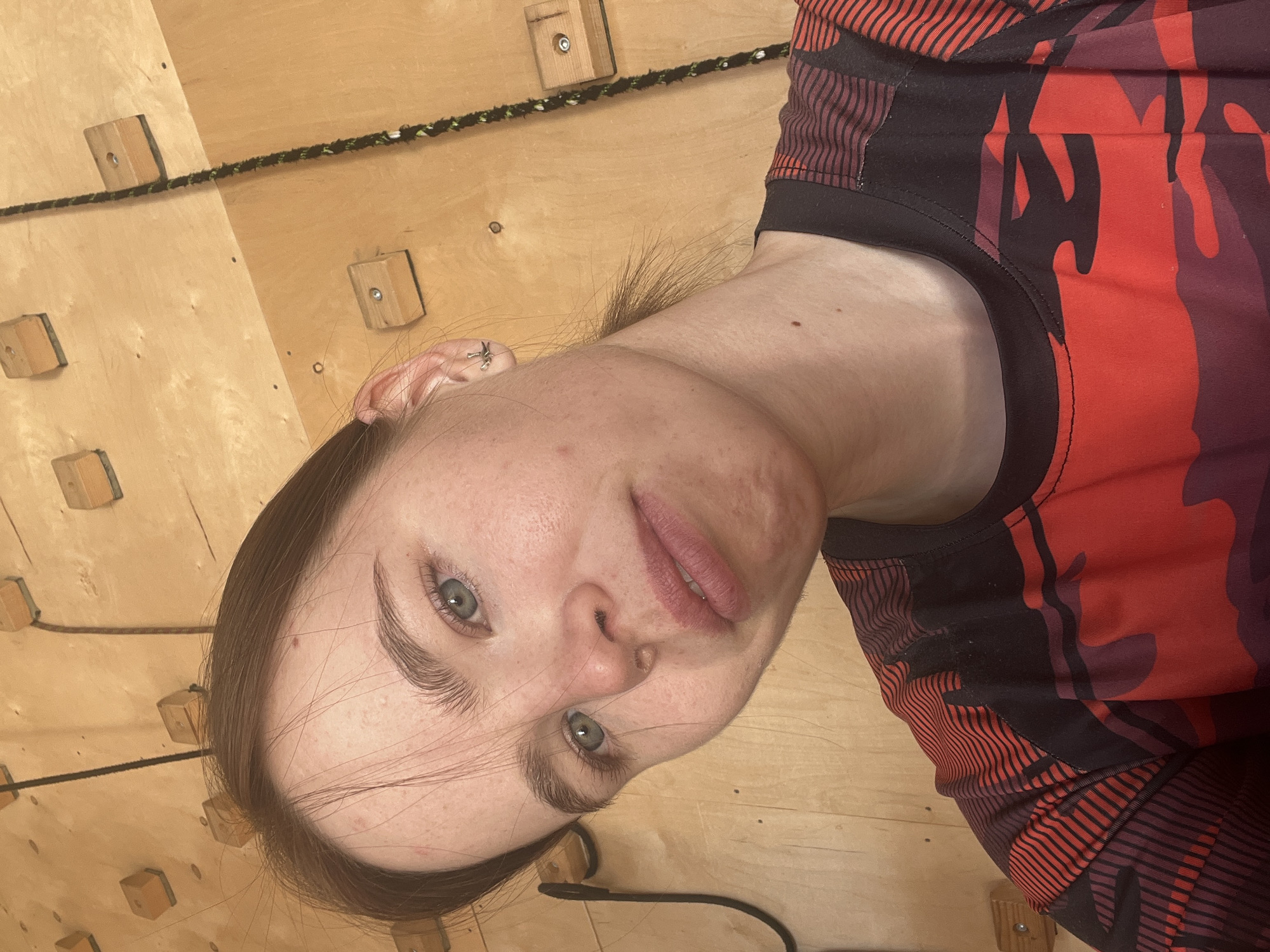 A woman is depicted in a selfie-style photograph. She is wearing a black and red camouflage-patterned top. She has light skin, brown hair, and green eyes. The background is a light wood wall with climbing holds attached. A black rope is also visible on the wall. There is no indication of the location being Chaikovsky, Russia in this image.