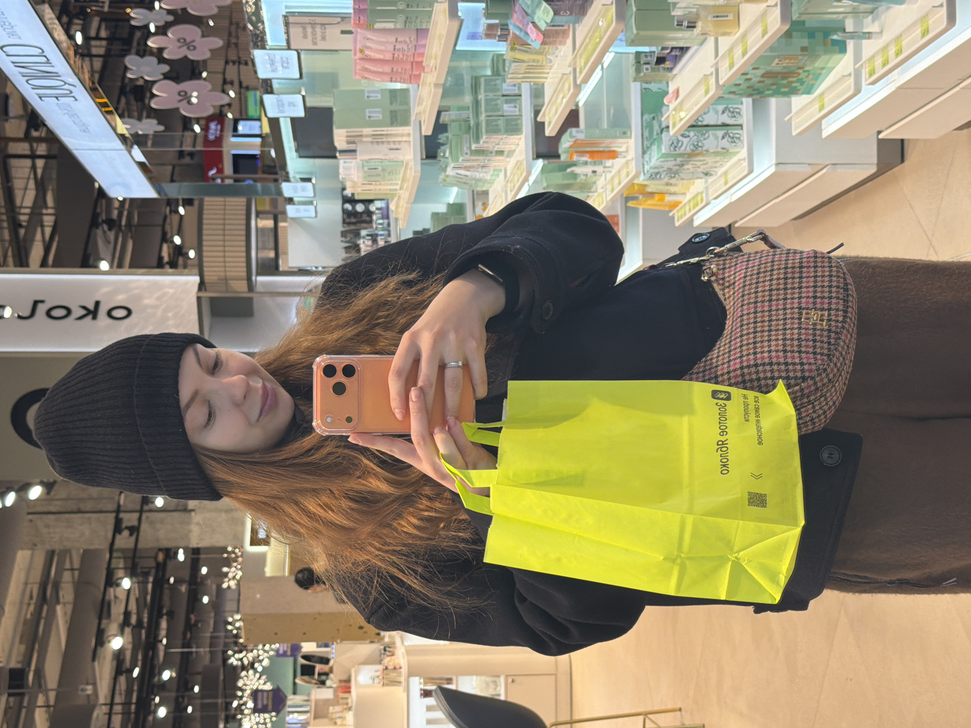 A young woman is depicted from the waist up, positioned within a retail store. She has long, reddish-brown hair and is wearing a black beanie, a black jacket or coat, and brown pants or a skirt. On her left hand, she wears a ring. She is holding an orange or rose gold colored smartphone with a clear case in both hands, appearing to interact with it, possibly taking a photo or viewing content. A small, plaid shoulder bag with a brown, pink, and dark checkered pattern and a gold-toned "EO" logo hangs on her right side. In her left hand, she also holds a vibrant neon yellow shopping bag.
The setting is a brightly lit cosmetics or beauty store. In the background, numerous shelves are visible, stocked with various small boxes and bottles, characteristic of beauty products. The ceiling features decorative white cloud-shaped cutouts, some adorned with a "%" symbol, suggesting a sale or promotional event. The store's interior has reflective surfaces, creating inversions and reflections of signs and lights.
The woman's expression is calm and appears content, with a slight smile. The lighting in the store is artificial and bright, suggesting an indoor setting where the time of day is not apparent.
Visible text on the neon yellow shopping bag reads "около ЗОЛОТОЕ ЯБЛОКО", which translates to "around GOLDEN APPLE," identifying the store as part of the "Golden Apple" cosmetics chain. Below this, the text "ВСЕ САМОЕ КЛАССНОЕ НА ЛЮБОЙ ДЕНЬ" (ALL THE COOLEST FOR ANY DAY) is also visible, along with a QR code. Reflected text on a shiny surface in the background includes "СТИЛЬНОЕ" (STYLISH).