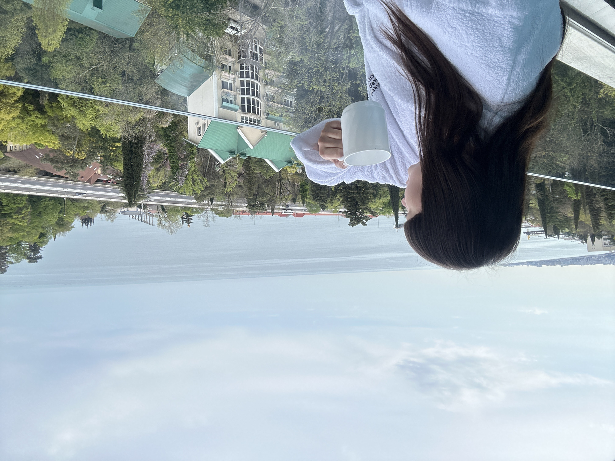 A woman in a white bathrobe holds a white mug and looks out over the water. The background features buildings and trees, indicating an urban setting. The image is in the city of Sochi, Russia. The environment consists of the sky, water, buildings, and trees. There is a road and some other buildings in the far background. The woman is the primary subject, and her action is observing the scenery while holding the mug.