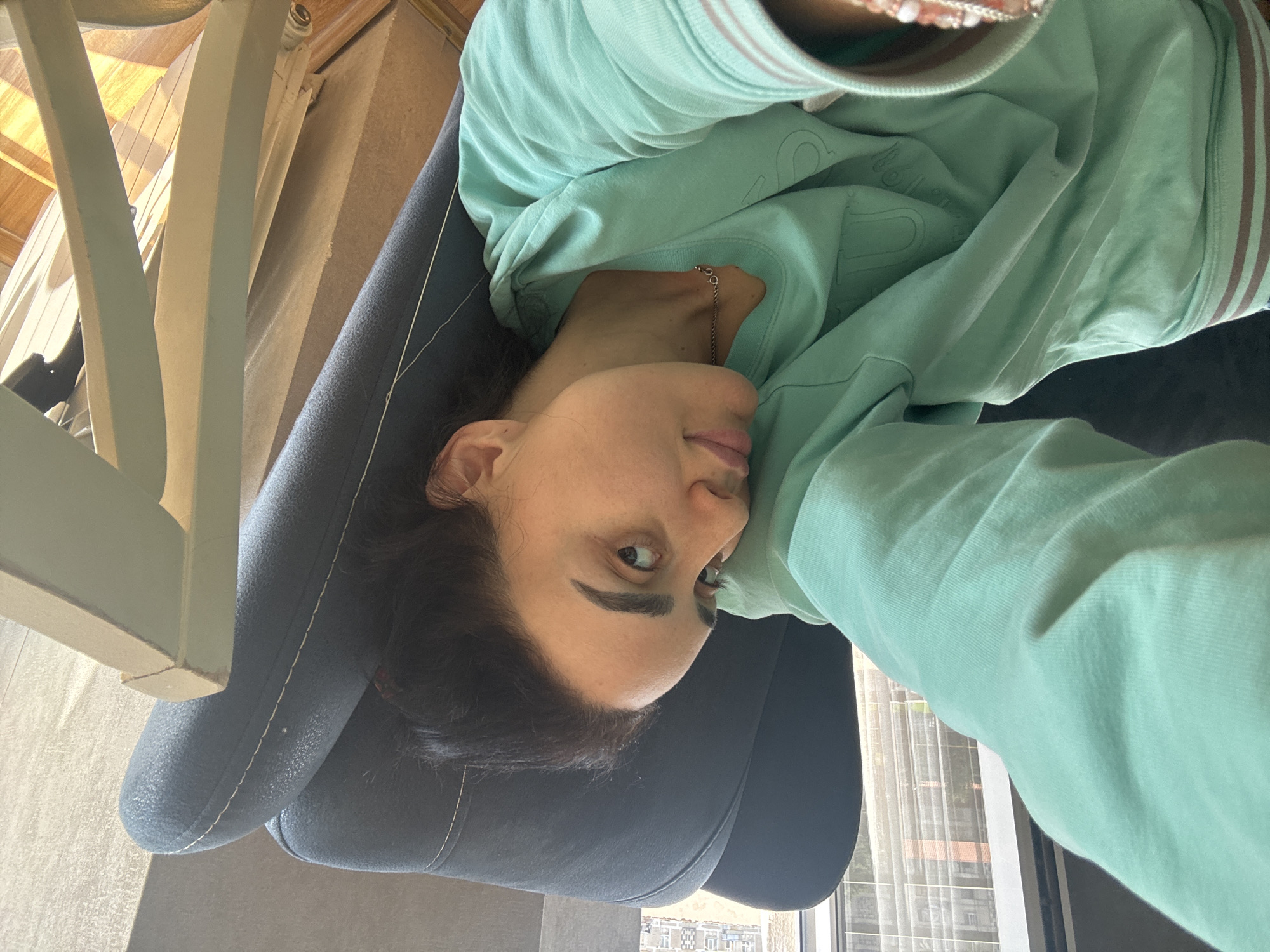 A woman in a mint-green sweatshirt is lying in a dark-blue chair. She is facing the camera. Her head is tilted to the left, and she has dark hair. A white table leg is in the top left corner of the image. A window with white curtains and a view of the city of Tbilisi, Georgia, is in the background. The buildings visible through the window appear to be residential.