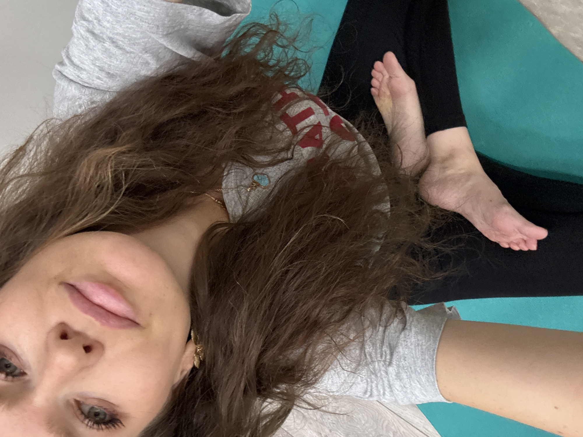 A woman lies on a blue yoga mat wearing black leggings and a gray t-shirt. She has long brown hair and is wearing a necklace. Her feet are visible on the mat. The background is a light gray wall and floor. The location is Unknown, Unknown.