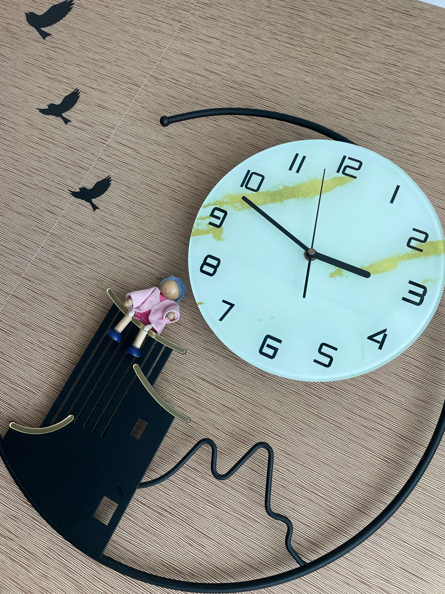 A clock wall decoration is laid flat on a textured brown surface. The clock face is round and pale blue with a faint yellow marbling. Black Arabic numerals mark the hours, and two black hands point to approximately 10:20. To the left of the clock is a black metal decorative element that resembles a stylized lighthouse or boat with a small wooden doll sitting on top. The doll has blue hair and is wearing a pink jacket and a darker pink dress. Three black silhouette birds are positioned in the upper left quadrant of the image, appearing to fly across the scene. The background surface has a woven texture with thin, parallel lines. The image is taken from a high-angle shot. There is no information available to indicate the location in the city of Pattaya City, Thailand.
