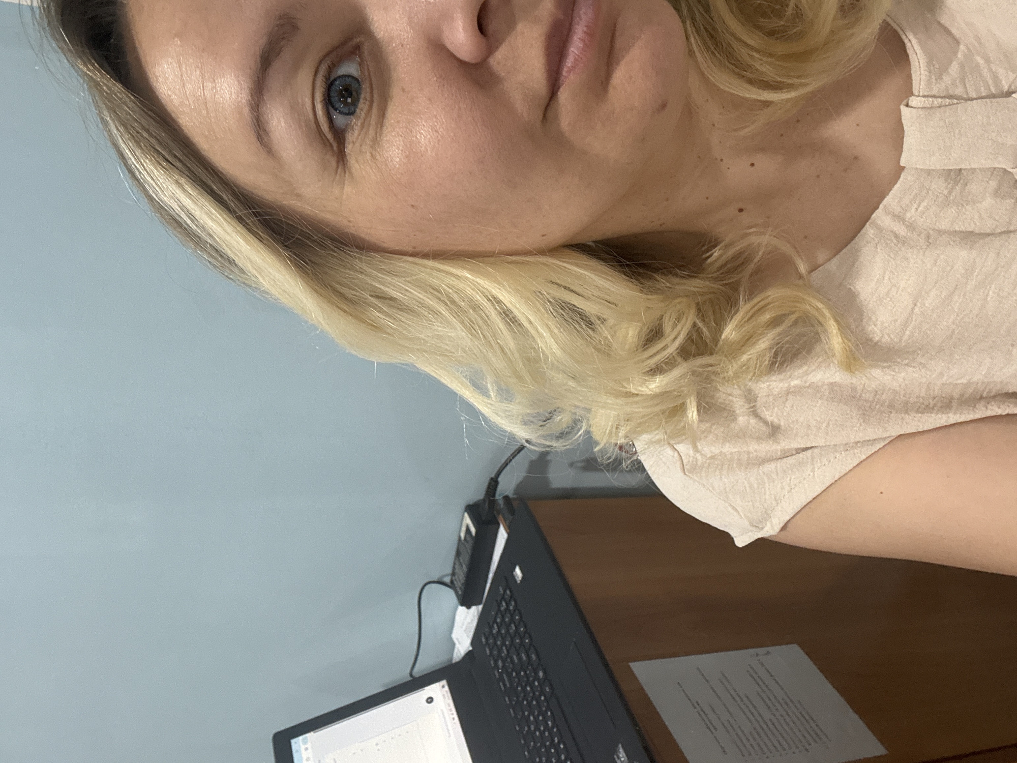 A woman with blonde hair and blue eyes is positioned in front of a laptop, which is on a wooden desk. A document rests next to the laptop. The background is a light blue wall. The location is not discernible as being in Beloretsk, Russia based on the image alone.