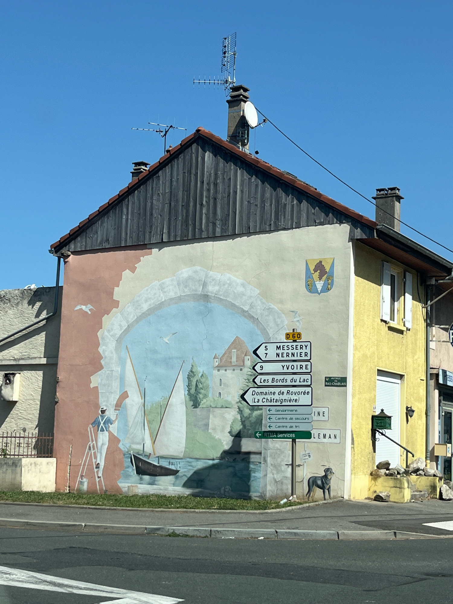 The image shows a mural painted on the side of a building in Douvaine, France. The mural depicts a harbor scene with sailboats, a castle, and a person painting the mural. There are also several road signs visible, indicating directions to Messery, Nernier, Yvoire, and other locations. A dog is painted standing near the base of the building. The building is yellow and appears to be a commercial establishment. The sky is clear and blue.