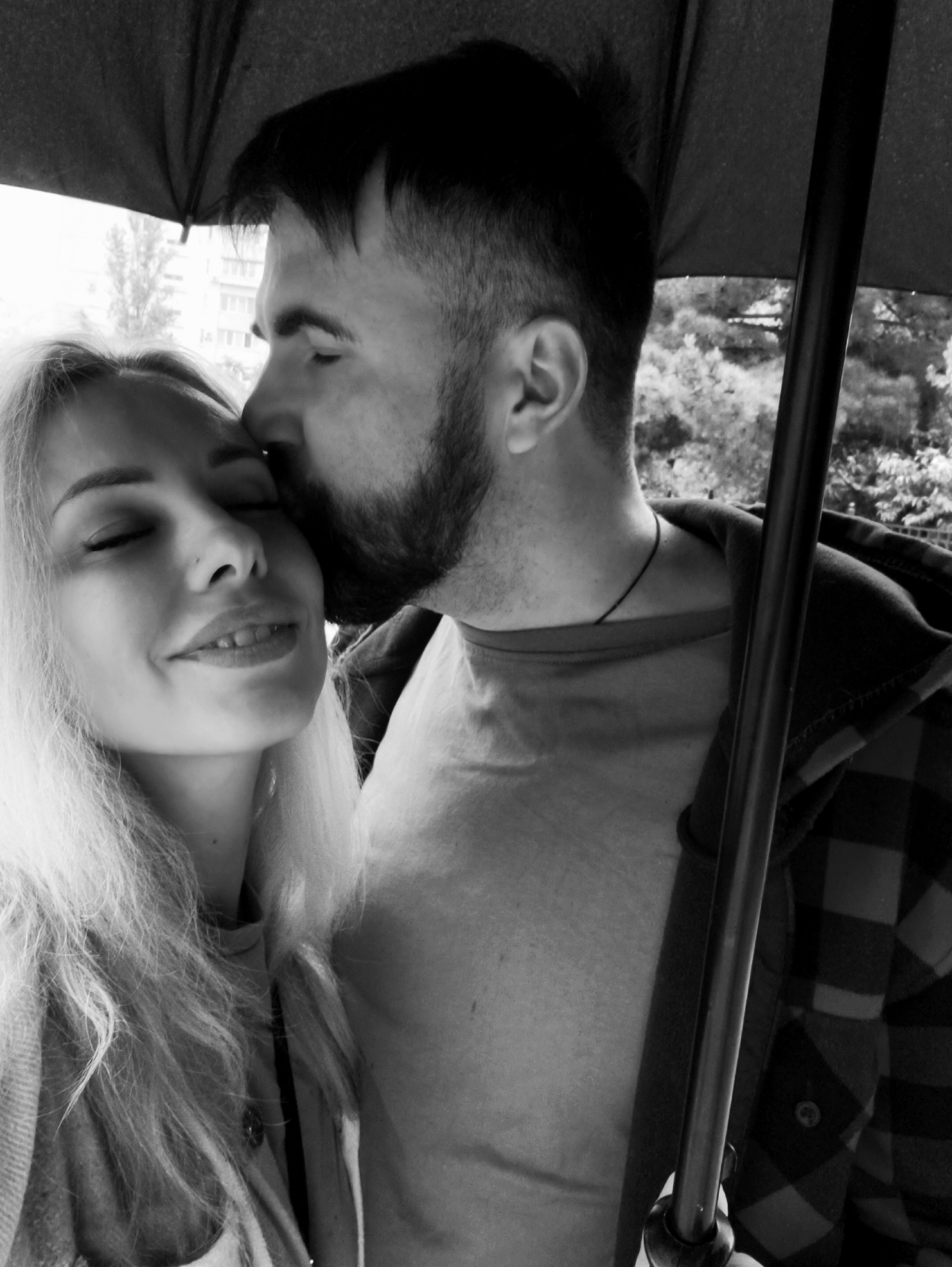 A black and white photograph depicts a couple under an umbrella. The man, with dark hair and a beard, is kissing the forehead of the woman, who has long blonde hair and closed eyes. The woman appears to be smiling. The man is wearing a t-shirt and a plaid jacket. The background shows trees and a building. The location is unspecified.