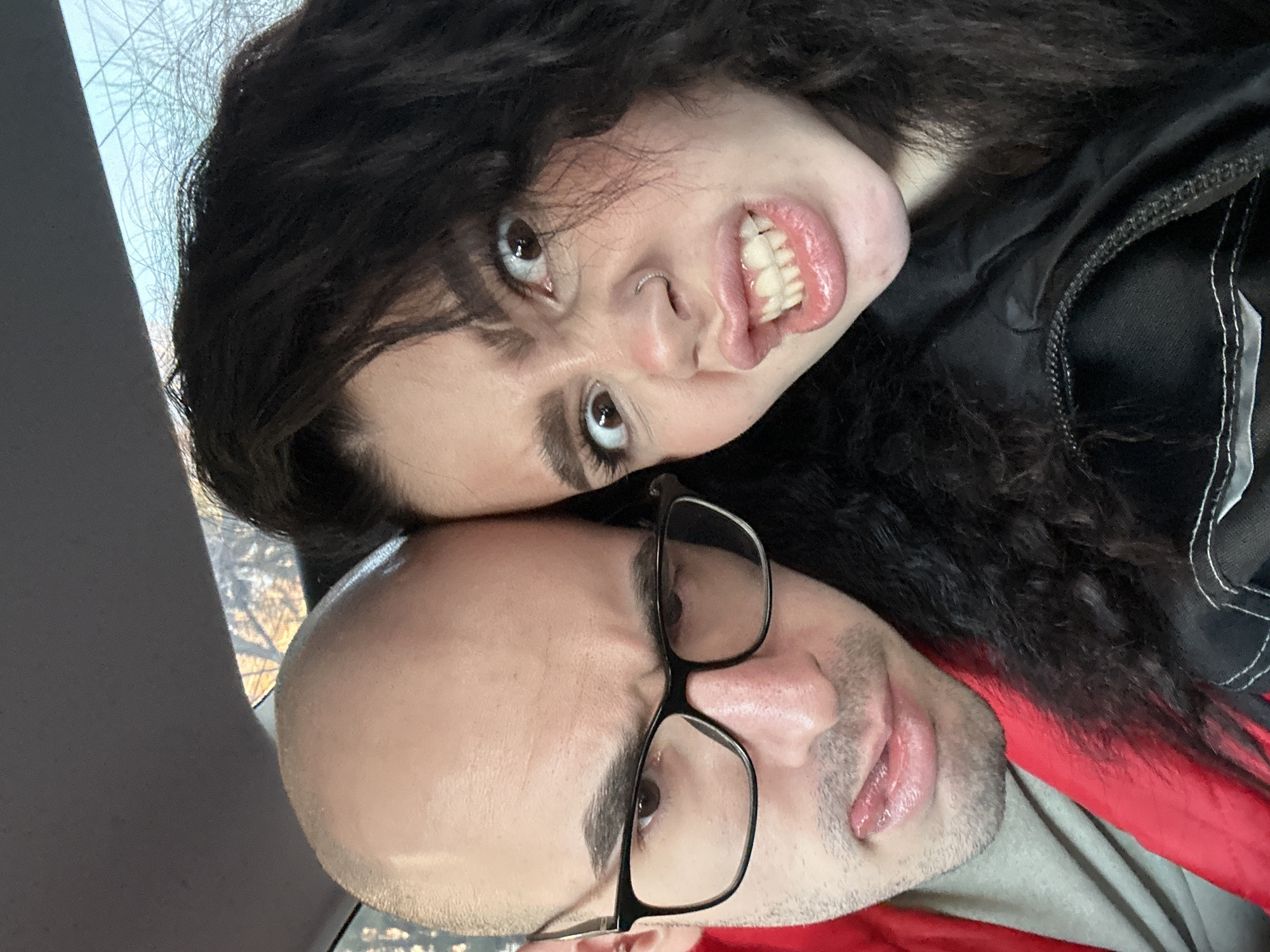 A photograph shows two people, one man and one woman, in a close-up shot, seemingly in an indoor environment with a window visible in the background. The woman has dark, curly hair and is positioned above the man. She is making a face with her mouth open, displaying her teeth, and her eyes are wide. The man is bald and wearing glasses, positioned below the woman. The background consists of what appears to be an interior space near a window, with some light coming through. The specific location in Kazan, Russia, is not discernible from the image.