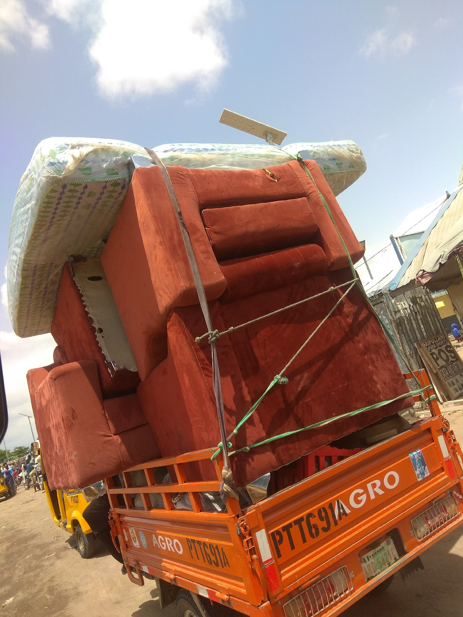 The image captures a bright, sunny day in Jalingo, Nigeria, with a clear blue sky and scattered white clouds. The scene is dominated by an orange truck heavily laden with furniture, primarily upholstered seats and mattresses. The furniture is stacked precariously high and secured with ropes.
In the foreground, the back of the orange truck is visible, featuring the text "AGRO PTT691A" in white and black lettering. There are also some stickers on the truck, including one with what appears to be a portrait. A license plate is also visible, reading "TARABA ARE 543 AH".
To the left of the truck, a yellow auto-rickshaw (Keke Napep) is partially visible, also loaded with items, and several people are seen around it, suggesting ongoing movement or a marketplace. In the background, there are market stalls with corrugated metal siding and awnings, indicating a bustling commercial area. One stall has a sign with "POS" prominently displayed. The overall impression is one of a busy, informal transportation and commerce scene in a Nigerian urban or semi-urban setting. The activity appears to be the loading or transport of goods, likely furniture, for sale or delivery.