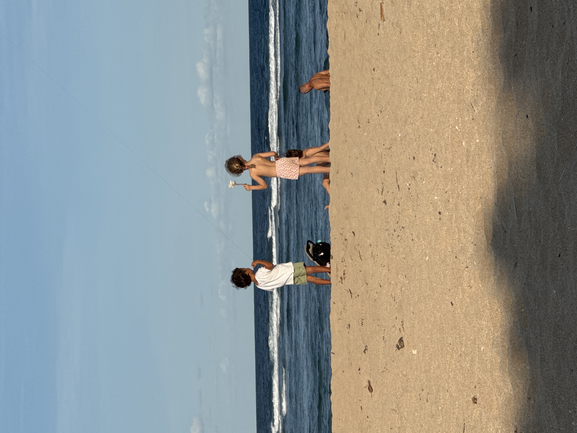 The image shows a beach scene in Kuta Selatan, Indonesia. The sky is blue with a few clouds. The ocean is blue with white waves. A child in a white shirt and green shorts is flying a kite. Two children are standing nearby, one in a pink swimsuit and the other covered by the first. A black bag is on the sand. Another person is partially visible in the background. The sand is tan and there are some shadows on it.