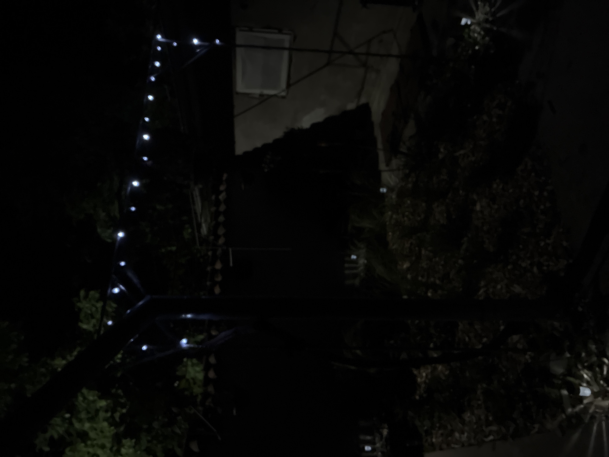 An outdoor scene at night features a string of white lights draped over a dark, angular structure. The lights illuminate a portion of the dark background, which includes trees and what appears to be a building with a window. The ground is covered in leaves, and there are faint glints of light from what may be ground-level solar lights. The overall impression is one of a dimly lit, possibly residential, outdoor space.