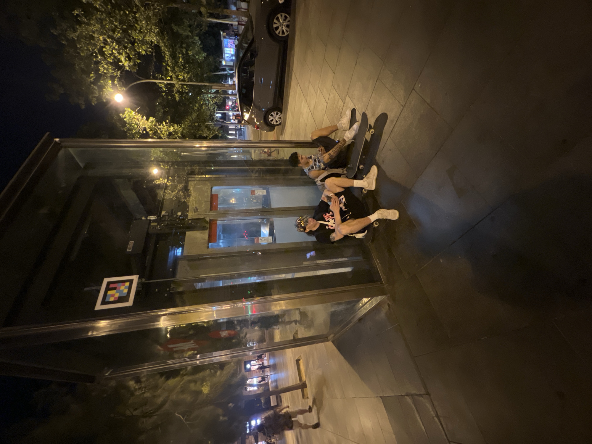 At nighttime on a city street in Barcelona, Spain, two young men sit on skateboards next to a glass enclosure that appears to be a bus stop or a modern phone booth. The enclosure is made of glass panels and metal framing, with reflections of the surrounding environment visible on its surfaces. One man, wearing a black t-shirt with a graphic design, a cap, and light-colored sneakers, sits with his legs extended and his skateboard beneath him. He is looking down at his phone. The second man, wearing a sleeveless patterned shirt and dark shorts, sits with his legs crossed, also looking at his phone, with his skateboard next to him. A gray SUV is parked on the street behind them. Trees with illuminated foliage line the sidewalk, and streetlights cast a warm glow on the scene. Pedestrians can be seen in the background, walking along the illuminated street and sidewalk. A colorful QR code-like marker is attached to the interior of the glass enclosure. The ground is paved with large, dark tiles.