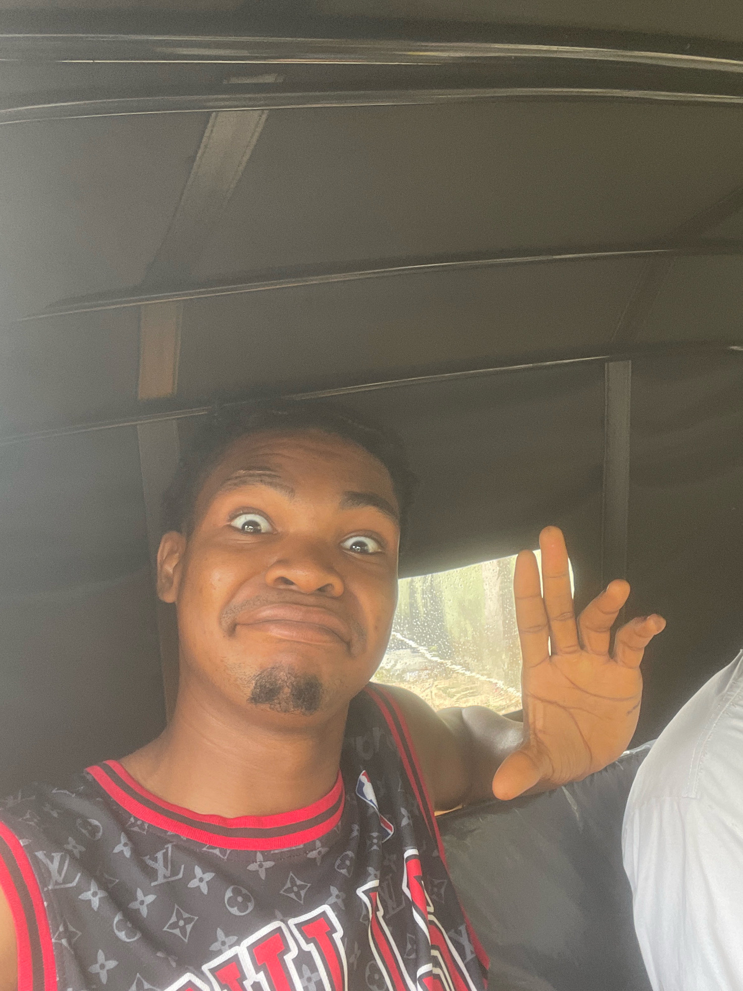 The image shows a young Black male in the foreground, appearing to be inside a vehicle. He is making a surprised or playful facial expression, with wide eyes and an open mouth. His right hand is raised, palm facing forward, with fingers slightly spread. He is wearing a black sleeveless jersey with a repeating Louis Vuitton pattern and red trim. A small goatee is visible on his chin.
Behind him, the interior of the vehicle appears to be dark, possibly with fabric lining the walls and ceiling. A wooden beam is visible above his head, suggesting the structure of the vehicle. To the right, the hand of another person, wearing a white garment, is visible. A window on the right side of the vehicle shows a blurred, bright outdoor scene, possibly indicating movement or a reflection.
While the specific location within Ado, Nigeria is not explicitly identifiable from the image alone, the context of being inside a vehicle that appears to be a tricycle or similar mode of transport is consistent with common transportation in Nigerian cities. The outdoor scene visible through the window offers a glimpse of the environment, likely a road or street with some greenery, though details are obscured by blurriness and potentially rain.