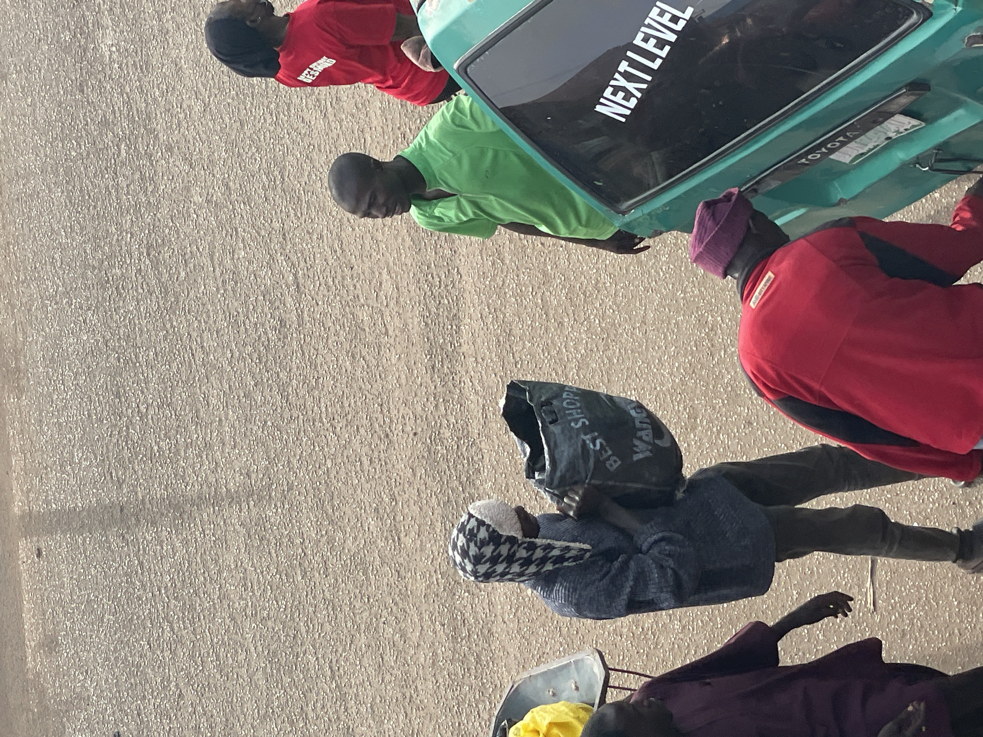 The image shows a group of people standing on a paved surface, possibly a street or parking lot, with a green vehicle in the background. The vehicle has "NEXT LEVEL" written in white on its windshield. The people are dressed in casual clothing. One person in the foreground is wearing a red shirt and appears to be looking down. Another person next to them is wearing a bright green shirt. A third person is wearing a red jacket and a purple beanie. In the lower part of the image, a person is wearing a dark blue hooded sweater and a patterned cap, holding a black bag with text on it. Another person is partially visible in a maroon garment at the bottom. The scene appears to be outdoors during the day, with the lighting suggesting it could be either morning or afternoon. The ground is a rough, textured pavement.