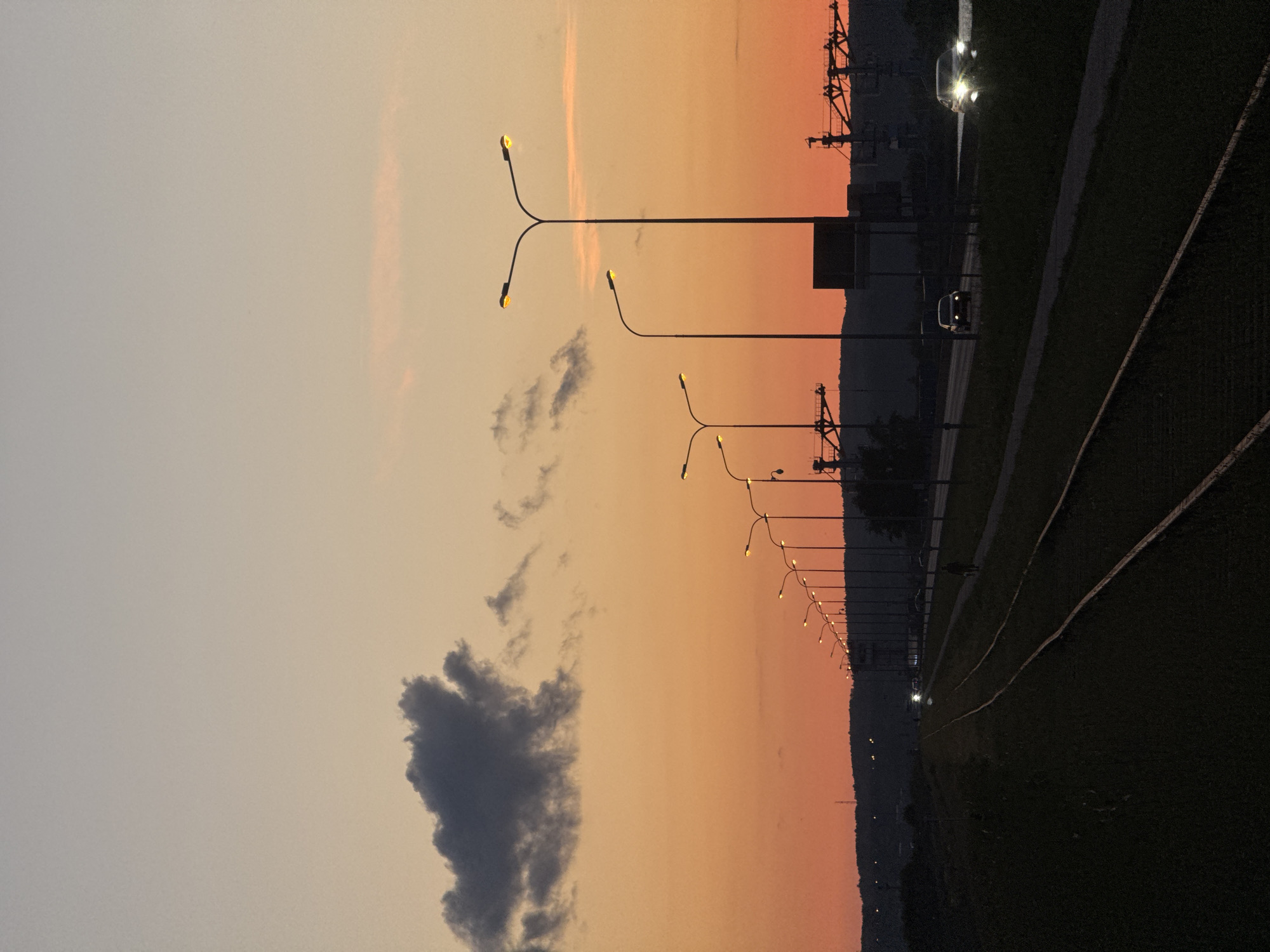 The image depicts a sunset scene with an orange sky. In the foreground, there are two sets of railroad tracks, dark and silhouetted. To the right of the tracks, a paved path runs parallel to a road where several cars are visible, some with headlights on. A line of streetlights illuminates the area along the road. In the background, there is a silhouette of a hill with some lights. Industrial cranes are visible on the right side of the frame. A single dark cloud hangs in the upper left portion of the sky. The scene appears to be located near the Kama River in Chaikovsky, Russia, given the presence of industrial structures often associated with port areas. There is a single pedestrian walking on the path.
