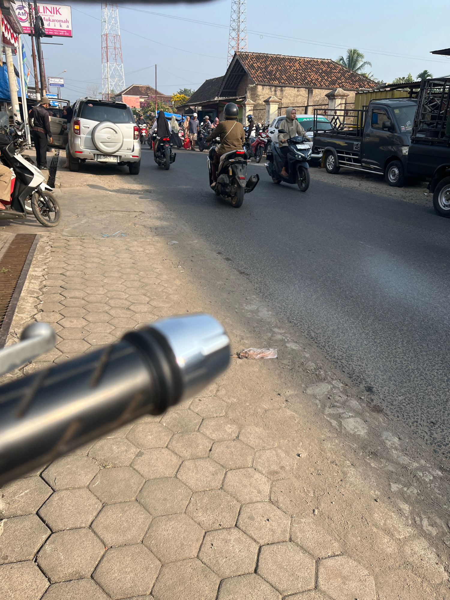 The image depicts a street scene in Kemuning, Indonesia, likely during the day due to the lighting. The foreground is dominated by the handlebars of a motorcycle, suggesting the image was taken from the perspective of a rider. The road surface appears to be asphalt, with a paved hexagonal tile sidewalk bordering it.
Various vehicles are visible on the road, including several motorcycles and at least two cars. One silver SUV with license plate "BE 3849 UD" is parked on the side of the road with its driver's side door open, and a person is standing next to it. Other motorcycles are in motion, with riders wearing helmets and casual clothing. A dark-colored pickup truck with a caged bed is also parked on the right side of the road.
Buildings line the street, some with traditional tiled roofs. A tall communication tower is visible in the background. Signs in Indonesian are present on the buildings, including one that reads "KLINIK AM INIK MUKAROMAH." The environment appears somewhat cluttered, with some litter visible on the side of the road. The overall impression is of a busy, ordinary street in a populated area.