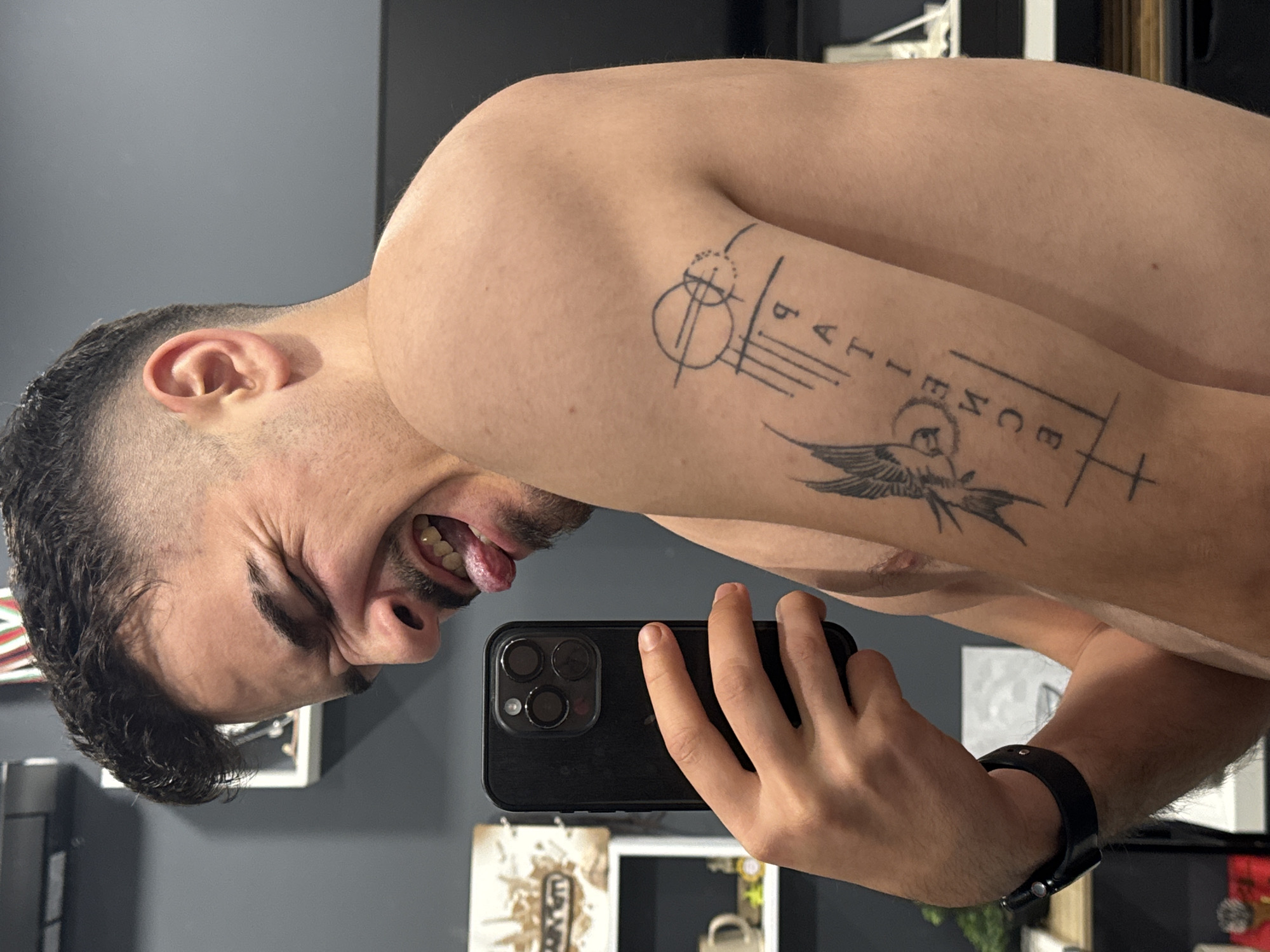 A man is taking a selfie in front of a mirror. He has short, dark hair, a mustache, and a tattoo on his upper arm that says "Patience" with other accompanying images. He is sticking his tongue out and grimacing. He is holding a black smartphone in his hand. He is wearing a black watch on his wrist. The background includes a gray wall, pictures hanging on the wall, and various objects on a shelf. The location is unknown and there is no indication that the picture was taken in Sofia, Bulgaria.
