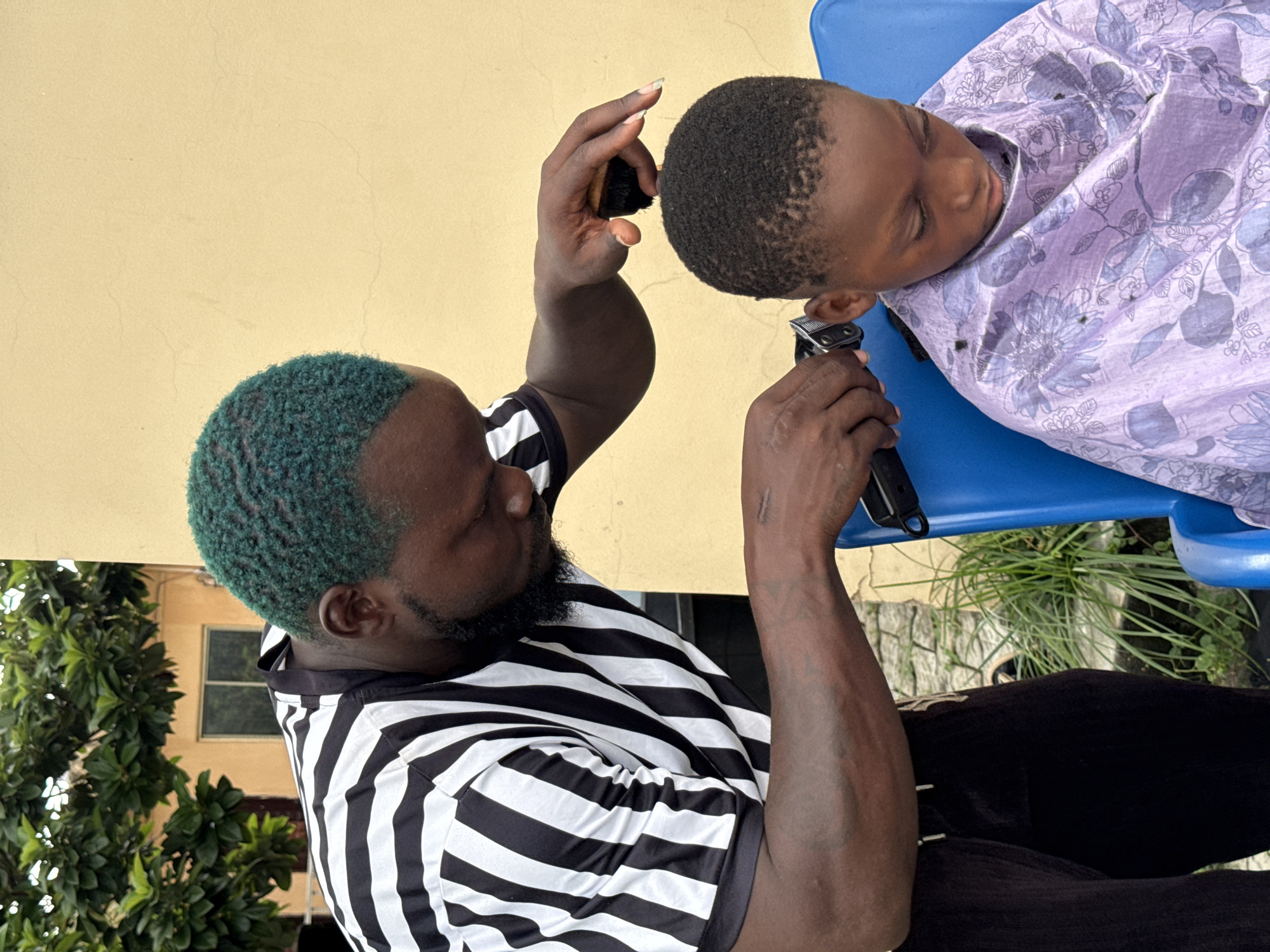 In Ado, Nigeria, a man with green-dyed hair, wearing a black and white striped shirt, is giving a haircut to a young boy. The boy is seated on a blue plastic chair, draped in a purple cape adorned with a floral pattern. The man is using electric clippers to cut the boy's hair, and he is also holding a small brush, possibly to sweep away loose hair. The scene appears to be outdoors, with a beige wall and lush green foliage visible in the background. The lighting suggests it is daytime.