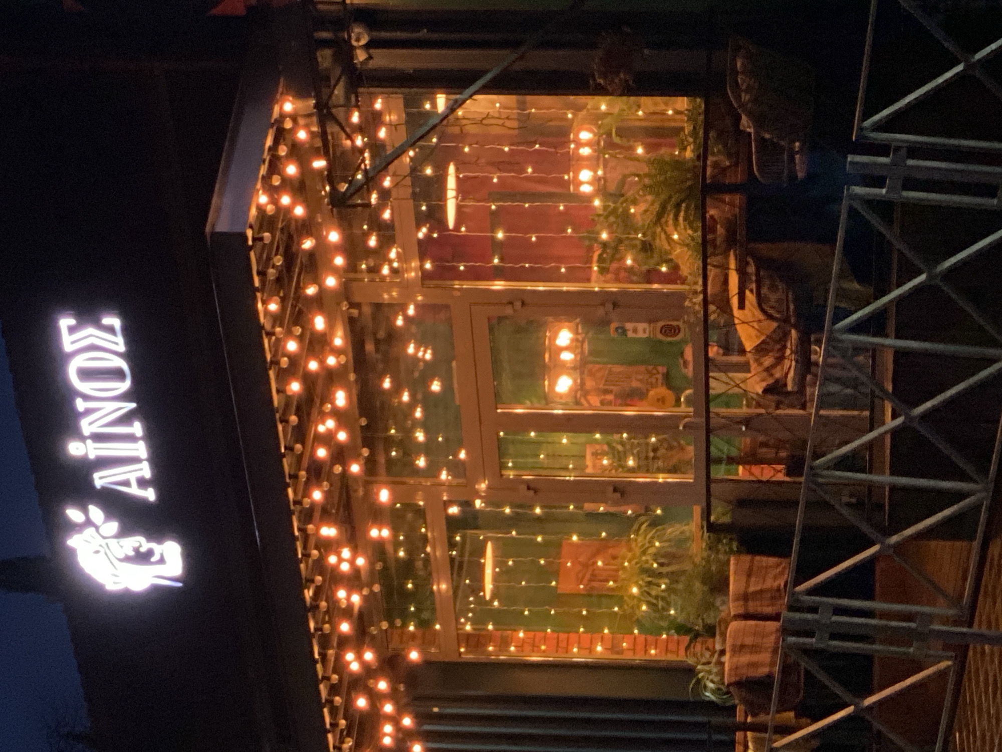 The image shows a nighttime view of the "AINOS" restaurant in Sochi, Russia. A lit sign displaying the restaurant's name vertically is visible on the left. The restaurant's exterior is adorned with numerous string lights. The facade is glass, allowing a view of the interior, which features plants and what appears to be brick accents. Metal scaffolding is partially visible on the right side of the image. No people are clearly visible in the shot.