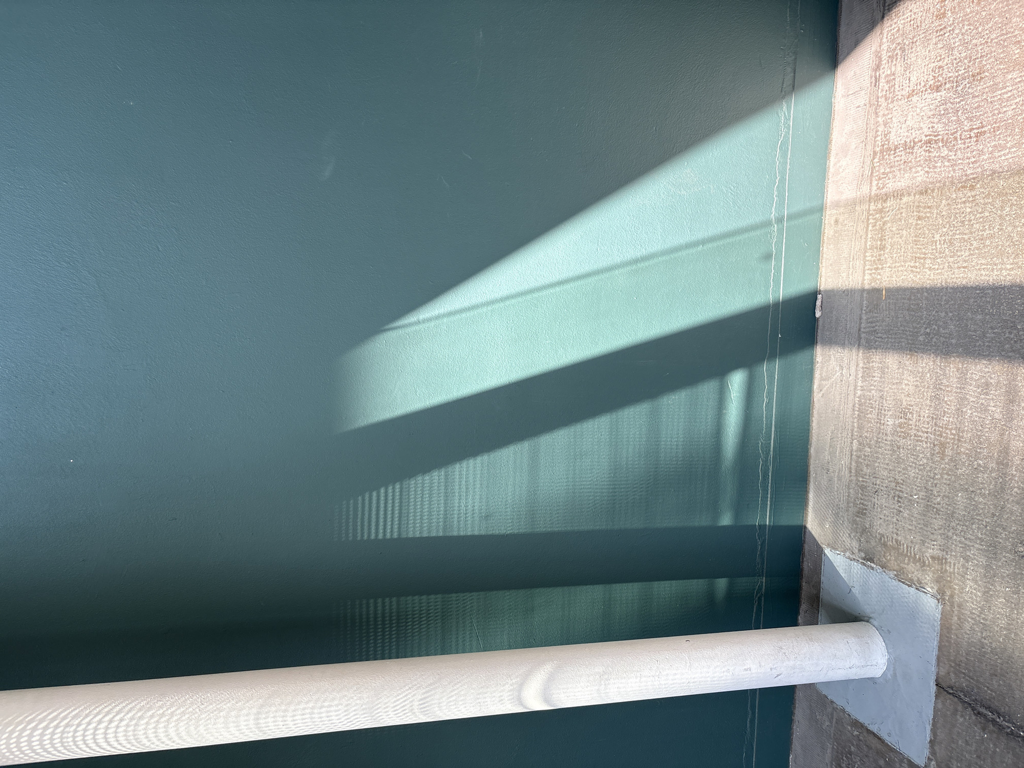 The image shows an indoor scene with a focus on a teal-colored wall, partially illuminated by sunlight creating distinct shadows. A white cylindrical pipe runs horizontally across the lower portion of the frame. The wall's texture is slightly rough. Adjacent to the teal wall is a rough, light-brown concrete surface. The sunlight casts elongated, angled shadows across both the teal wall and the pipe. There are thin vertical drips or lines on the teal wall near its edge. The pipe appears to be sealed or joined to the concrete wall with a patch of grey sealant.