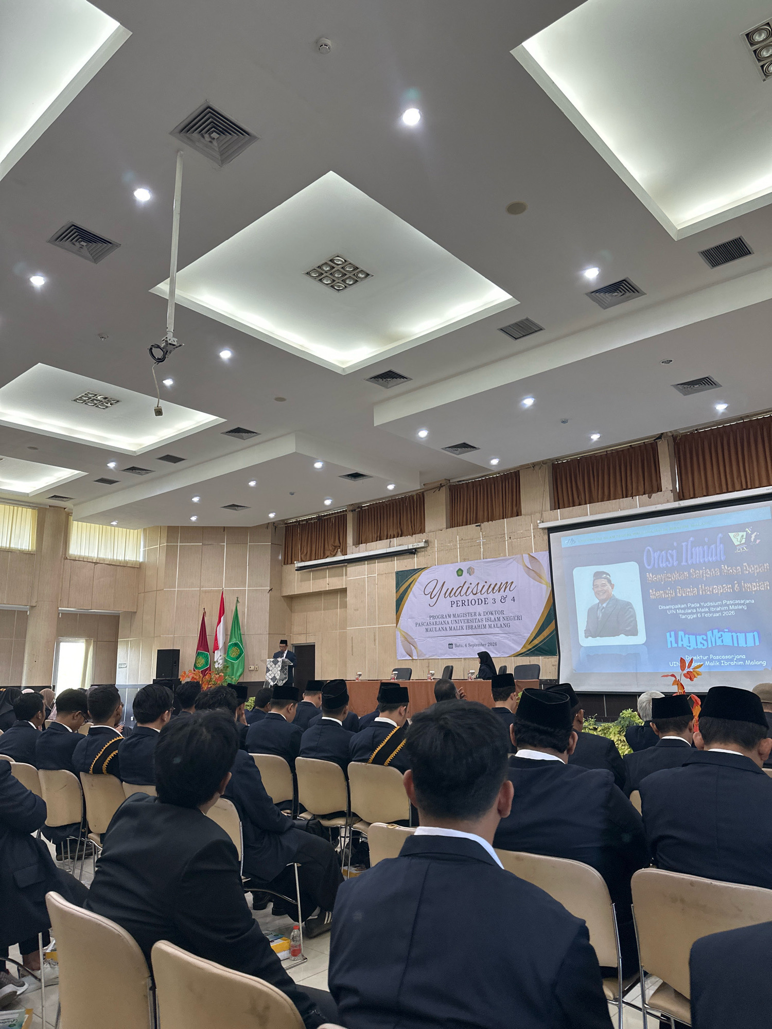 The image depicts an indoor gathering, likely a commencement or academic ceremony, held within the city of Batu, Indonesia. The scene is set in a large hall with a high, coffered ceiling featuring recessed lighting and air vents. The audience, composed primarily of men wearing black suits and traditional Indonesian peci hats, is seated in rows of chairs, facing a stage area.
The stage is adorned with two flags, one red and white (the Indonesian national flag) and another green with an emblem, likely representing an institution. In front of the flags, a speaker stands at a podium, delivering an address. To the right of the stage, a large projection screen displays an image of a man in a suit and peci, accompanied by text in Indonesian. One banner on the left side of the stage reads "Yudisium PERIODE 3 & 4 PROGRAM MAGISTER & DOKTOR PASCASARJANA UNIVERSITAS ISLAM NEGERI MAULANA MALIK IBRAHIM MALANG" and below it "Batu, 4 September 2020". Another banner to the right of the screen shows a similar emblem and text which appears to be related to the event. The overall atmosphere suggests a formal academic event.