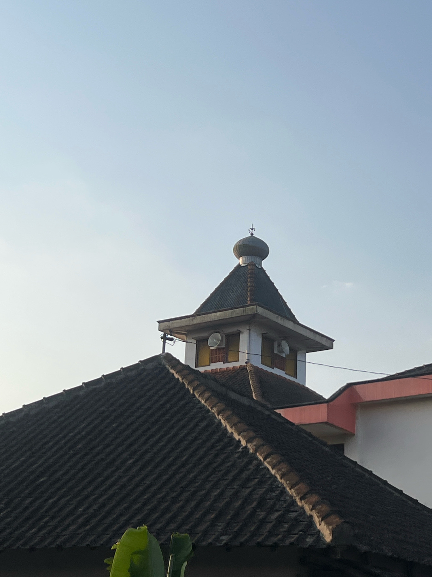 The image shows the rooftop of a building in Madyocondro, Indonesia, against a clear sky. The foreground features a dark, tiled roof with a prominent ridge. Behind this, a multi-tiered structure rises, topped by a dome. This upper section of the building is painted white with yellow accents around windows. Two white circular objects, possibly speakers or air conditioning units, are visible on this level. A small metallic finial adorns the very top of the dome. To the right, a portion of another building with a pink and white facade is visible, also with a tiled roof. The scene is static, with no people or discernable activity. The lighting suggests it is daytime, possibly morning or late afternoon due to the soft light.