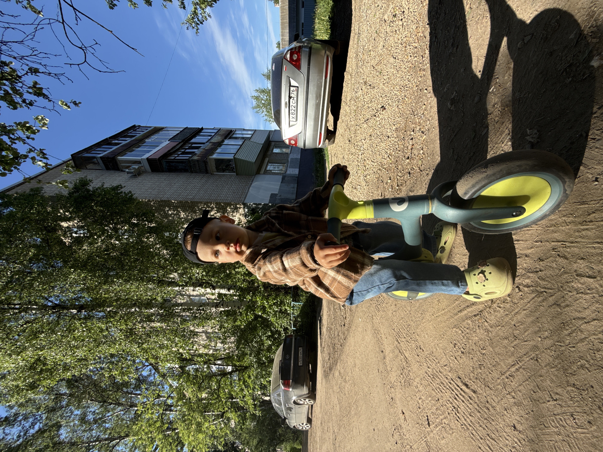 A young boy wearing a brown plaid shirt, blue jeans, a black cap, and green Crocs is sitting on a green and yellow tricycle on a gravel path. The location is Rusanovo, Russia as evidenced by the car license plates that show the Russian flag and country code “RUS”. To the left are trees and a multi-story brick building with visible balconies. A silver sedan with the license plate X822 CM 76 and a black sedan with the license plate H530 KM 76 are parked adjacent to the path. The sky is blue with light clouds. The scene is captured in bright sunlight, casting shadows of the bike and boy onto the ground.