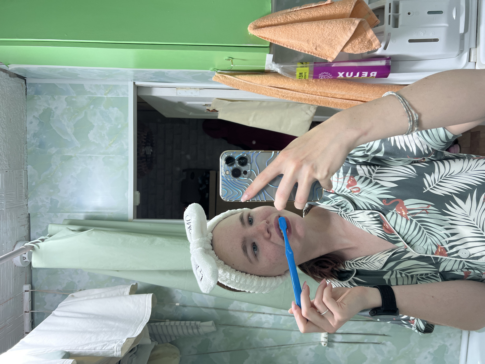 In a bathroom with light green patterned walls, a young woman stands in front of a mirror, brushing her teeth with a blue toothbrush. She is wearing pajamas with a tropical leaf and flamingo print and a white fuzzy headband with a bow. Her phone, with a blue and silver wave-patterned case, is held in her right hand as she takes a selfie. In the background, laundry hangs on a clothesline, and orange towels are draped over hooks. A tube of "BETOX" cream is visible on a shelf. The scene appears to be in a residential bathroom.