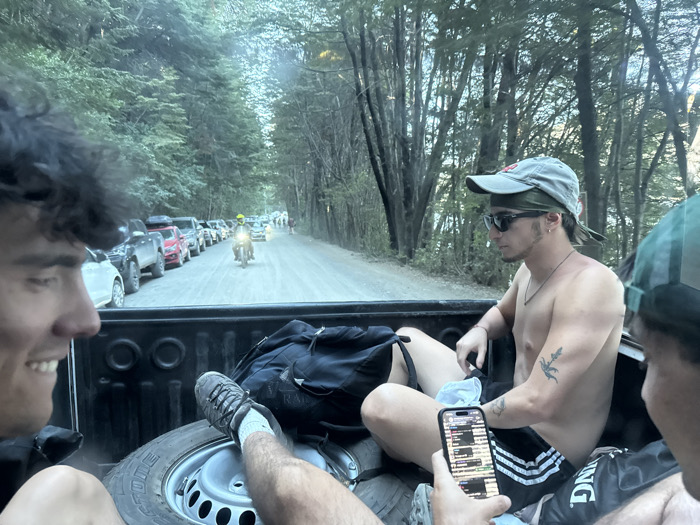 Three shirtless young men ride in the back of a pickup truck. One is looking at his phone, another is smiling, and the third is partially visible. A spare tire and backpack lie in the truck bed. The truck drives on a dirt road lined with trees and parked cars. A motorcycle follows behind. The scene suggests an outdoor adventure.
