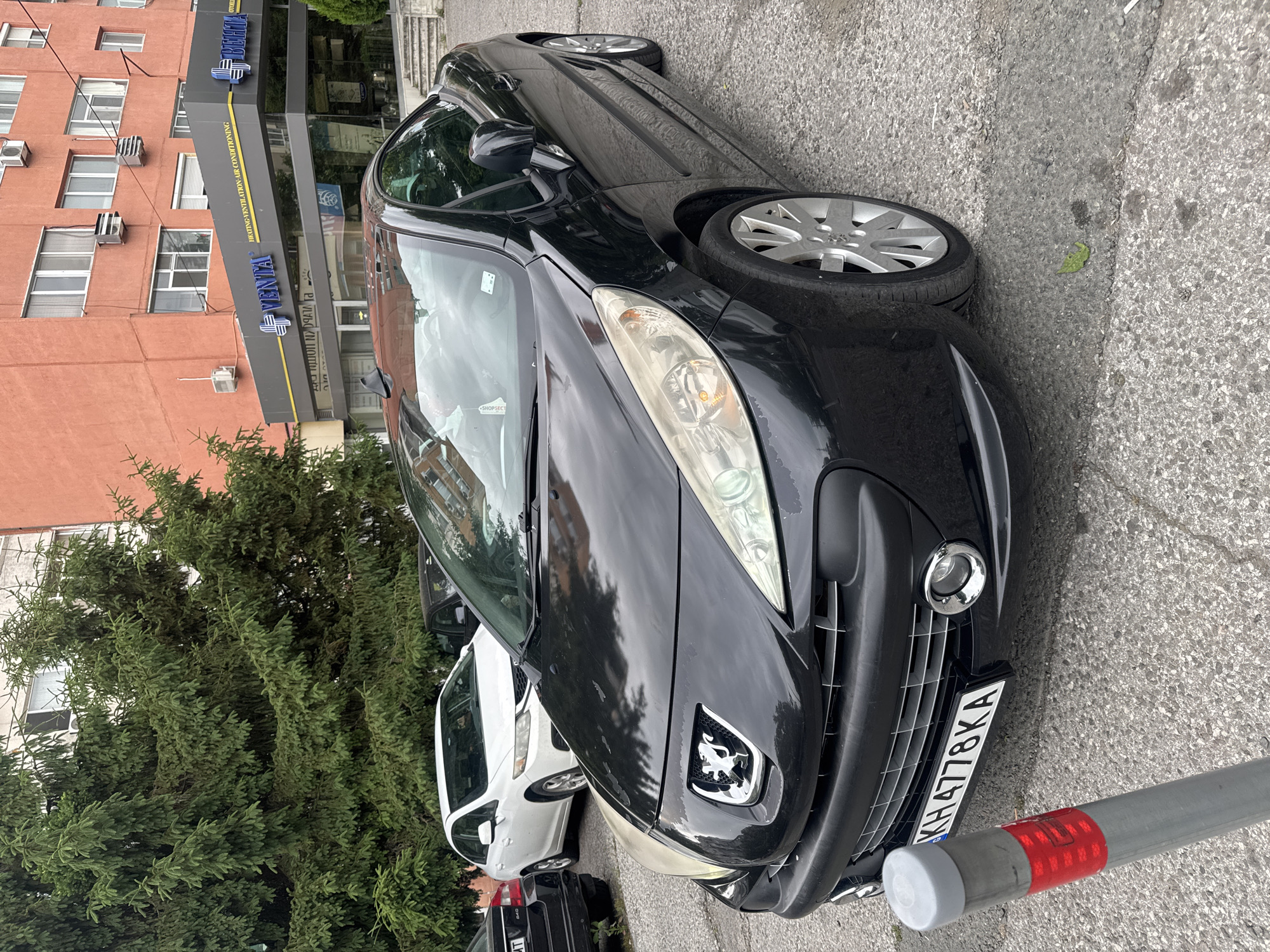A black Peugeot 207 CC convertible is parked on a paved surface in front of a building. The car's front end is facing the viewer, with its license plate clearly visible as KH4778KA. The building behind the car is a multi-story structure with an orange facade, and a sign above the ground floor entrance reads "VENTA HEATING-VENTILATION-AIR CONDITIONING". Several large evergreen trees are situated to the left of the car. Other vehicles, including a white sedan and a dark-colored car, are partially visible in the background. A metal bollard with reflective red markings stands to the left of the Peugeot. The overall scene appears to be an outdoor parking area in an urban environment.