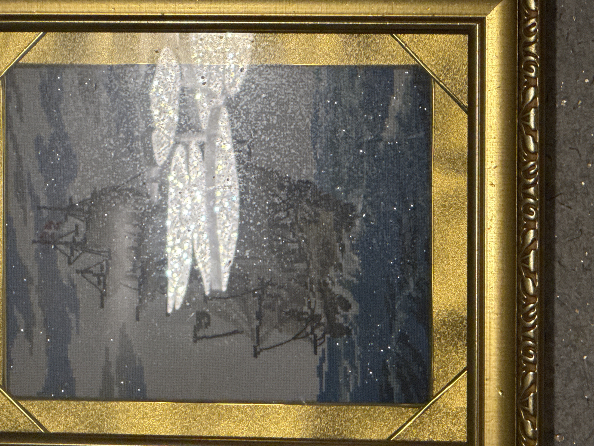 This is a framed embroidered picture of a tall ship sailing on a choppy sea. The ship is depicted in dark tones, with its masts and rigging clearly visible. The sea is rendered in shades of blue and grey, with white crests of waves splashing against the ship. There are numerous small, white specks scattered across the image, which appear to be glitter or some other reflective material, possibly intended to represent sea spray or light on the water. The frame is ornate and golden, with a textured surface and decorative molding at the bottom. The background of the embroidery is a pale grey sky with darker clouds. The scene appears to be a stormy or rough sea condition. The image is a close-up, with the bottom edge of the frame and a textured, dark surface below it visible. There is no indication of any specific location within the city of Ulaanbaatar, Mongolia, as the image depicts a maritime scene.