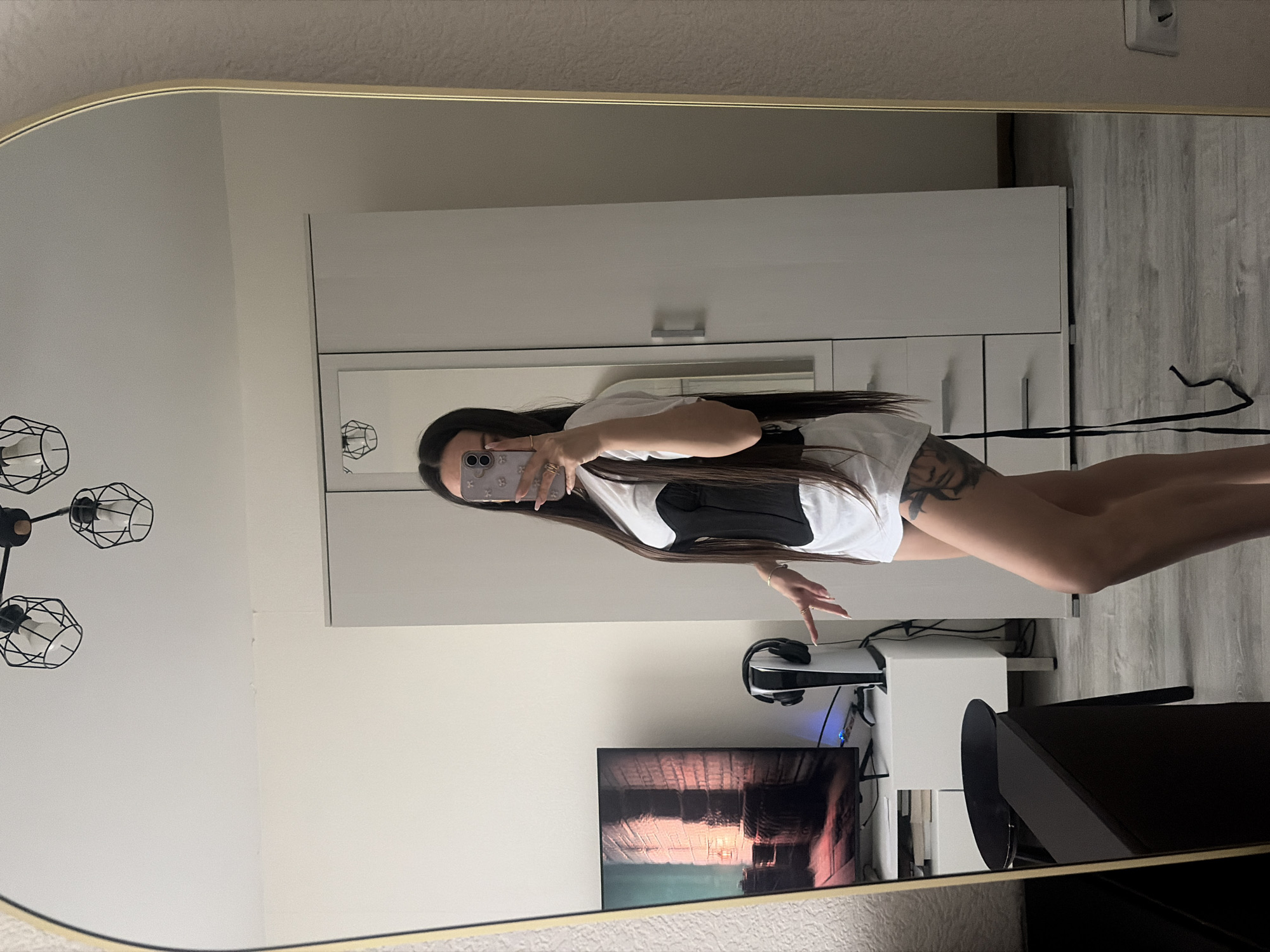 A mirror reflects a woman in a white t-shirt and black bustier, holding a phone, against a backdrop of a white wardrobe, gaming setup with a PS5, and a TV showing an abstract image. The scene appears to be indoors, potentially a bedroom or living area, in a modern, well-lit setting. There is no indication of the location being in Saint Petersburg, Russia.
