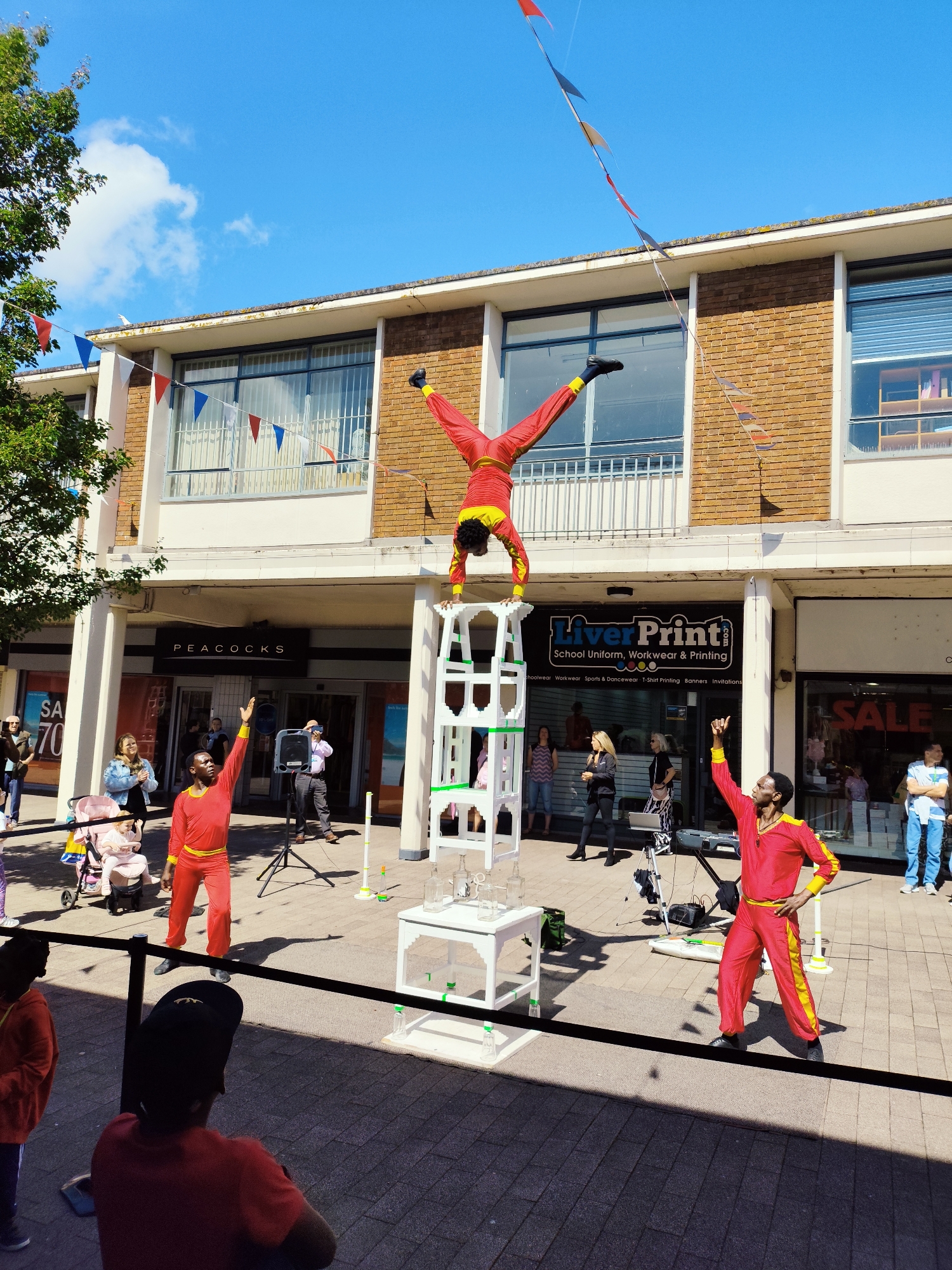 This image depicts a street performance taking place in a pedestrianized area outside a row of shops. The architecture suggests a modern urban setting, possibly a shopping precinct. The shops include "PEACOCKS" and "LiverPrint" which advertises "School Uniform, Workwear & Printing". A banner indicating "SALE 70" is visible on another shopfront. The ground is paved with dark grey and lighter grey paving stones. A black rope barrier runs across the foreground.

The main focus is on a street performance. In the center of the image, a performer in a red and yellow costume is balancing upside down on a stack of white tiered structures. Another performer, also in a red and yellow costume, stands to the left, facing the audience and with one arm raised. A third performer in a similar costume is to the right, also with an arm raised, striking a pose. The performers appear to be part of a circus or acrobatic troupe.

In the background, several spectators are visible, including a woman pushing a pink stroller with a baby in it. A person holding a microphone stands near a speaker system on a tripod. Other individuals are seen standing and observing the performance.

The sky is blue with some white clouds. Red, white, and blue bunting is strung across the area, suggesting a festive occasion or public event. Trees are visible on the left side of the image, indicating the presence of some greenery in the urban environment. The overall scene is brightly lit by sunlight, casting distinct shadows on the paved ground. The location is identified as Knowsley, United Kingdom.