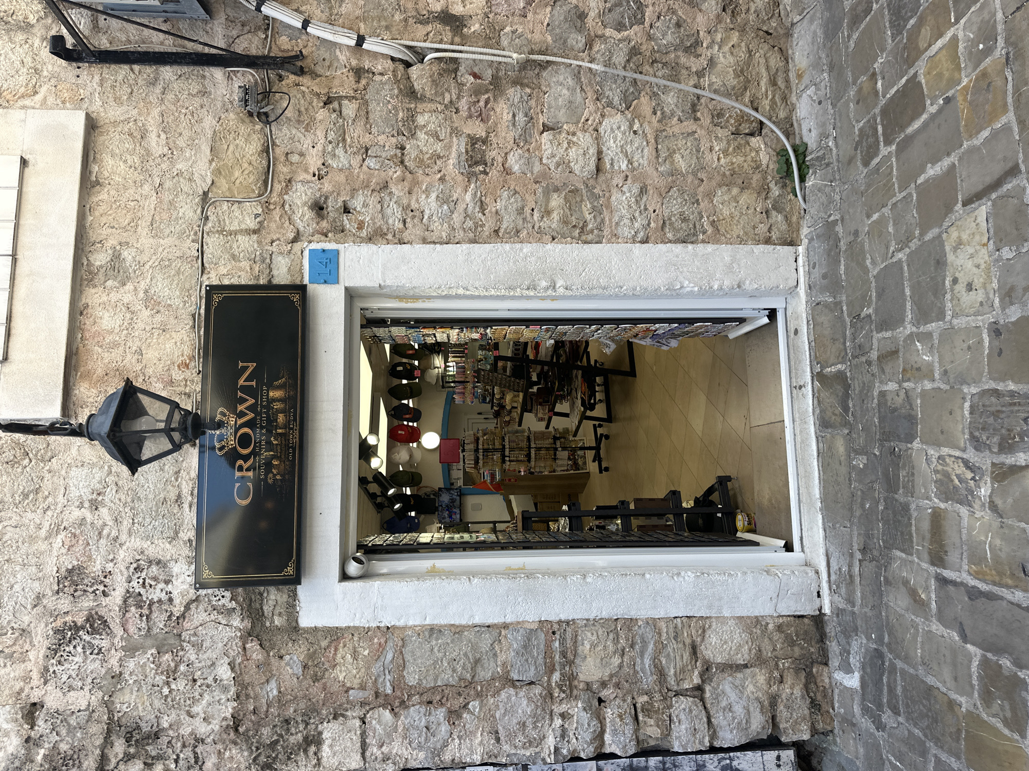 The image shows the storefront of a souvenir and gift shop called "Crown" in the Old Town of Budva, Montenegro. The shop's entrance is framed by a white stone archway set into a wall constructed of light-colored stone blocks of varying sizes. To the left of the doorway is a black sign with gold lettering that reads "Crown HANDMADE SOUVENIRS & GIFT SHOP OLD TOWN BUDVA" and is mounted below a black metal lamppost. The interior of the shop is visible through the doorway, revealing racks of merchandise including hats, jewelry, and other souvenirs, displayed on shelves and tables within the brightly lit space. The floor appears to be tiled. Electrical wiring and conduit run along the wall above the entrance. The wall to the right of the doorway is constructed of grey stone blocks.