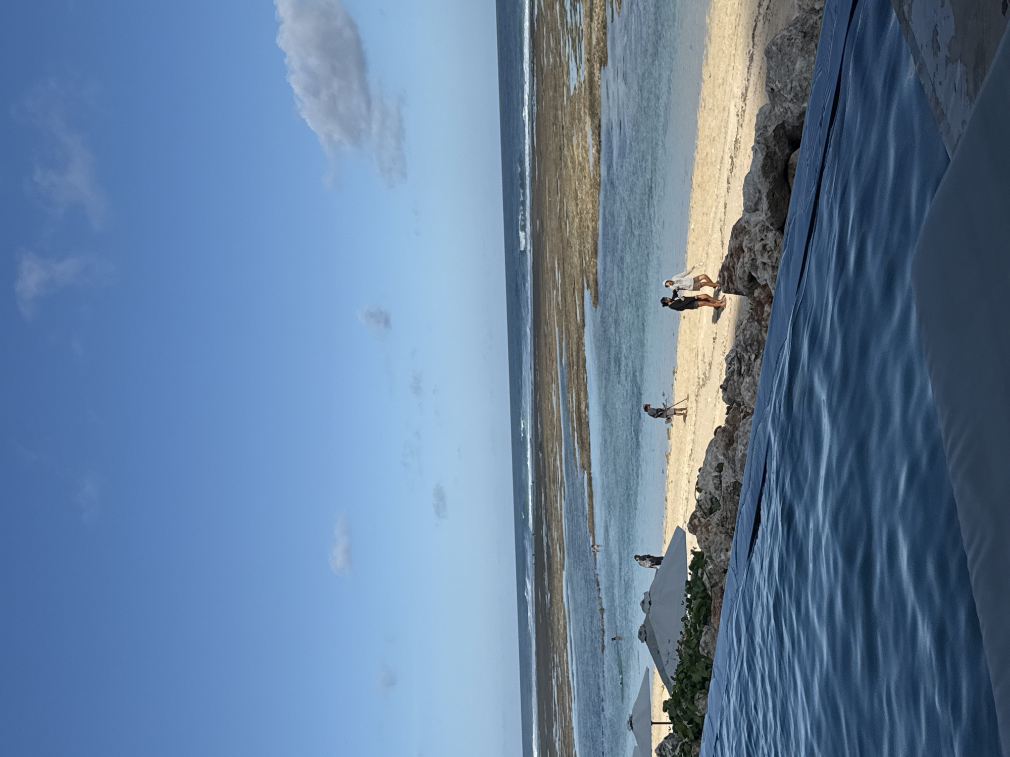 This image captures a beach scene in Kuta Selatan, Indonesia. The sky is clear blue with a few scattered clouds. The shoreline consists of sand and exposed rocks or coral, transitioning into the ocean. The tide appears to be low, revealing a wide expanse of shallow water with what looks like a coral reef structure. There are people on the beach, some standing near the rocks, others walking along the shoreline, and one individual farther out in the shallow water. There are also white umbrellas on the shoreline closer to the right. A pool or body of water is visible in the foreground, rippling and obscuring some of the beach.