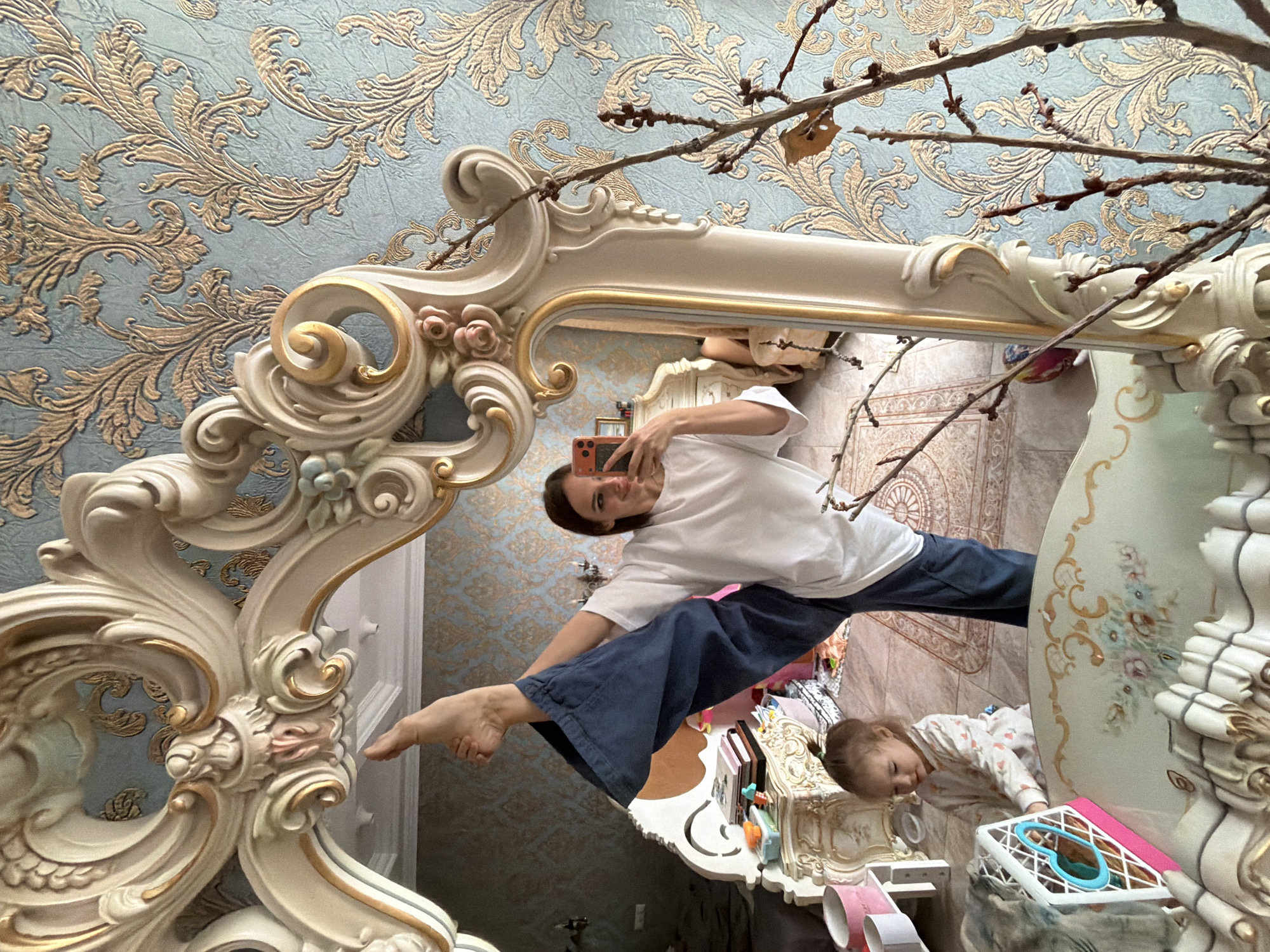 In Ufa, Russia, a woman is taking a mirror selfie in a room. She is performing a split, with one leg extended high in the air. She is wearing a white t-shirt and blue pants. A young child is sitting on the floor to her left, playing with a toy. The room is decorated with ornate furniture and patterned wallpaper. A large, decorative mirror is prominently featured in the image, reflecting the scene.