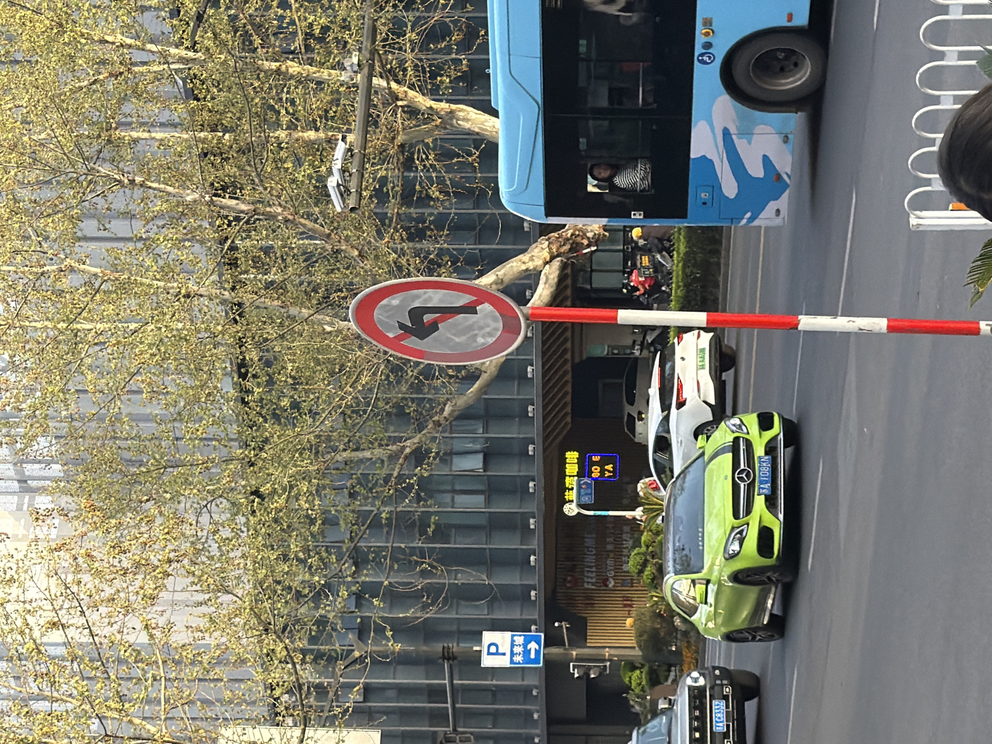 The image captures a street scene in Xuanwu District, China, with a "no left turn" traffic sign prominently displayed against a backdrop of an office building and a tree. A light blue bus passes in the background, and a lime green Mercedes Benz is parked on the right side of the frame. Other cars and motorcycles are visible on the street. A parking sign with an arrow pointing upwards is mounted on a pole. Signs displaying Chinese characters, including a reference to "Lanwan Cafe," are also visible.