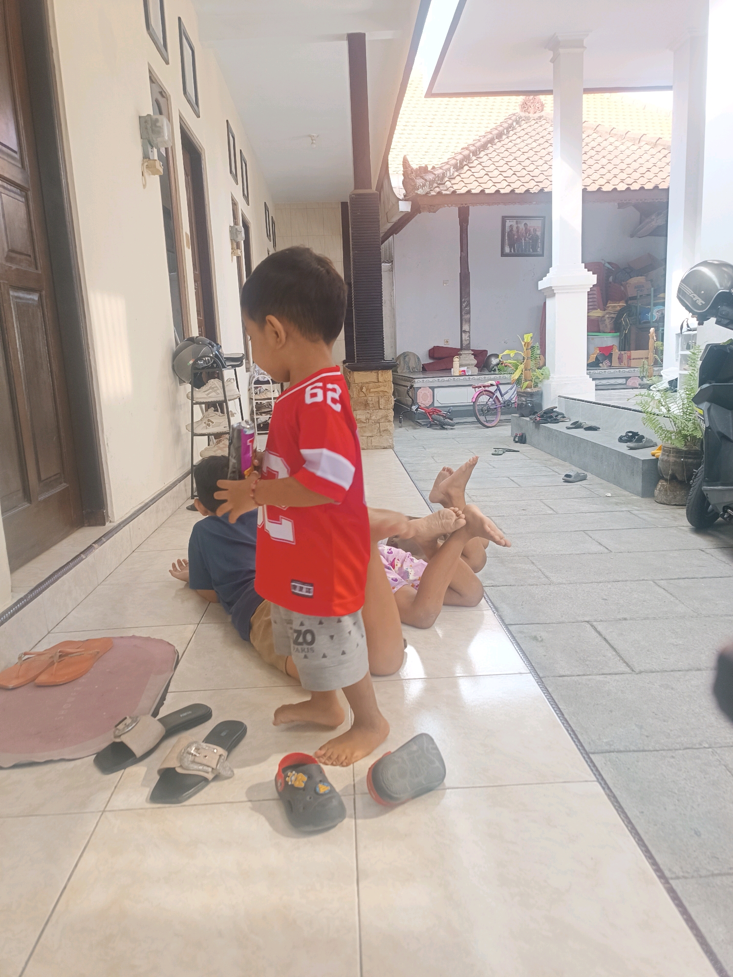The image depicts a scene at a residence in Tabanan, Indonesia. In the foreground, a young boy wearing a red shirt with white sleeves and grey shorts stands facing away from the camera, looking towards the left. He is holding a purple object. To his right, another young child, possibly a girl based on the pink and white patterned shorts and pink top, is lying on their back on the tiled floor, with their legs raised. The boy in the red shirt appears to be interacting with the child on the ground, as his right hand is on the other child's head.
The environment appears to be an outdoor or semi-outdoor area of a house, characterized by tiled flooring and architectural elements consistent with Indonesian housing, such as roof tiles and a covered patio area. Several pairs of footwear, including flip-flops and slip-on sandals, are scattered on the tiles in the foreground and midground. In the background, a motorcycle helmet, a bicycle, and various stored items are visible, suggesting a lived-in space. The lighting indicates it is daytime.