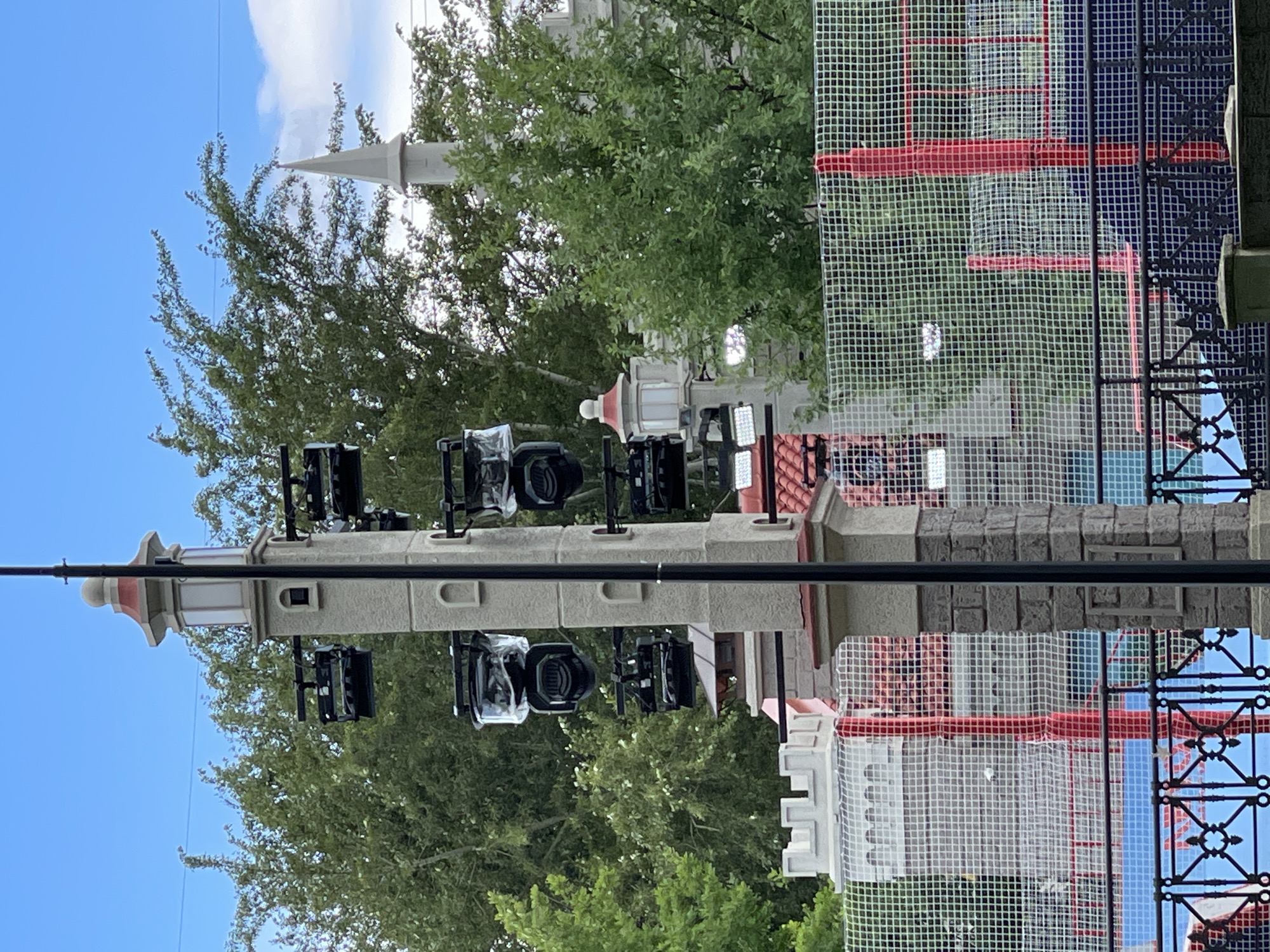 This media file shows a vertical shot of an outdoor scene, possibly a public square or park, with a castle-like structure in the background and lush green trees. A tall, ornate light post is prominently featured in the foreground, adorned with several professional-looking spotlights. Behind the light post, a section of a building with a red and white checkered facade and a net-like covering is visible. The sky is blue with some white clouds, suggesting fair weather. The presence of the spotlights and the net-covered structure could indicate preparations for an event, a performance, or ongoing construction/renovation work. The overall atmosphere is neutral, with no discernible emotions or specific activities being captured. No visible text is present in the image. Given the "Location context: Moscow, Russia" prompt, it's plausible this scene is located in Moscow.