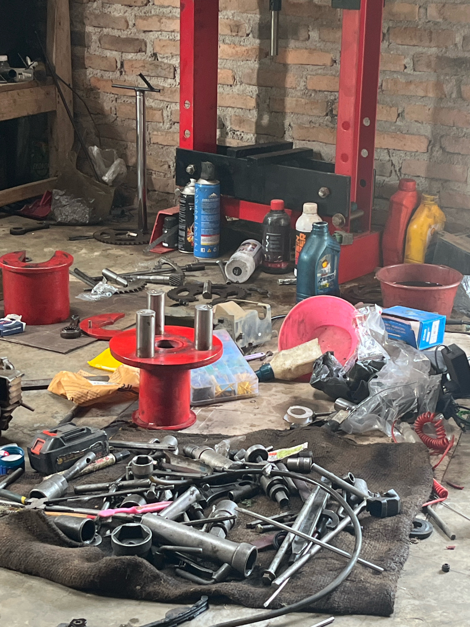 The image depicts a cluttered workshop area, likely in Kebumen, Indonesia, characterized by a brick wall in the background and a concrete floor covered with tools and various items. Dominating the center is a red hydraulic press. Scattered on the floor are numerous tools including wrenches, sockets, pliers, and a battery-powered drill. Red and yellow plastic containers, along with several bottles of liquids, are also visible. A large brown tarp lies on the floor, piled with more tools. The overall impression is one of an active, but disorganized, workspace.