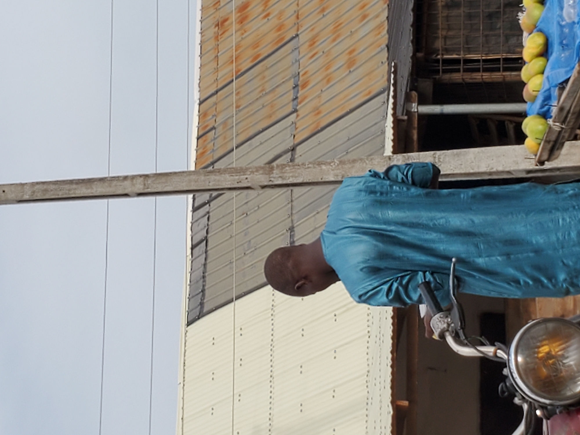 A man is standing next to a motorcycle with a headlight visible in Mararaba, Nigeria. He is wearing a blue garment. The motorcycle appears to be carrying a load of fruit covered with blue material. In the background, there is a building with a corrugated metal exterior, some of which shows rust. Overhead electrical lines are also visible in the sky.