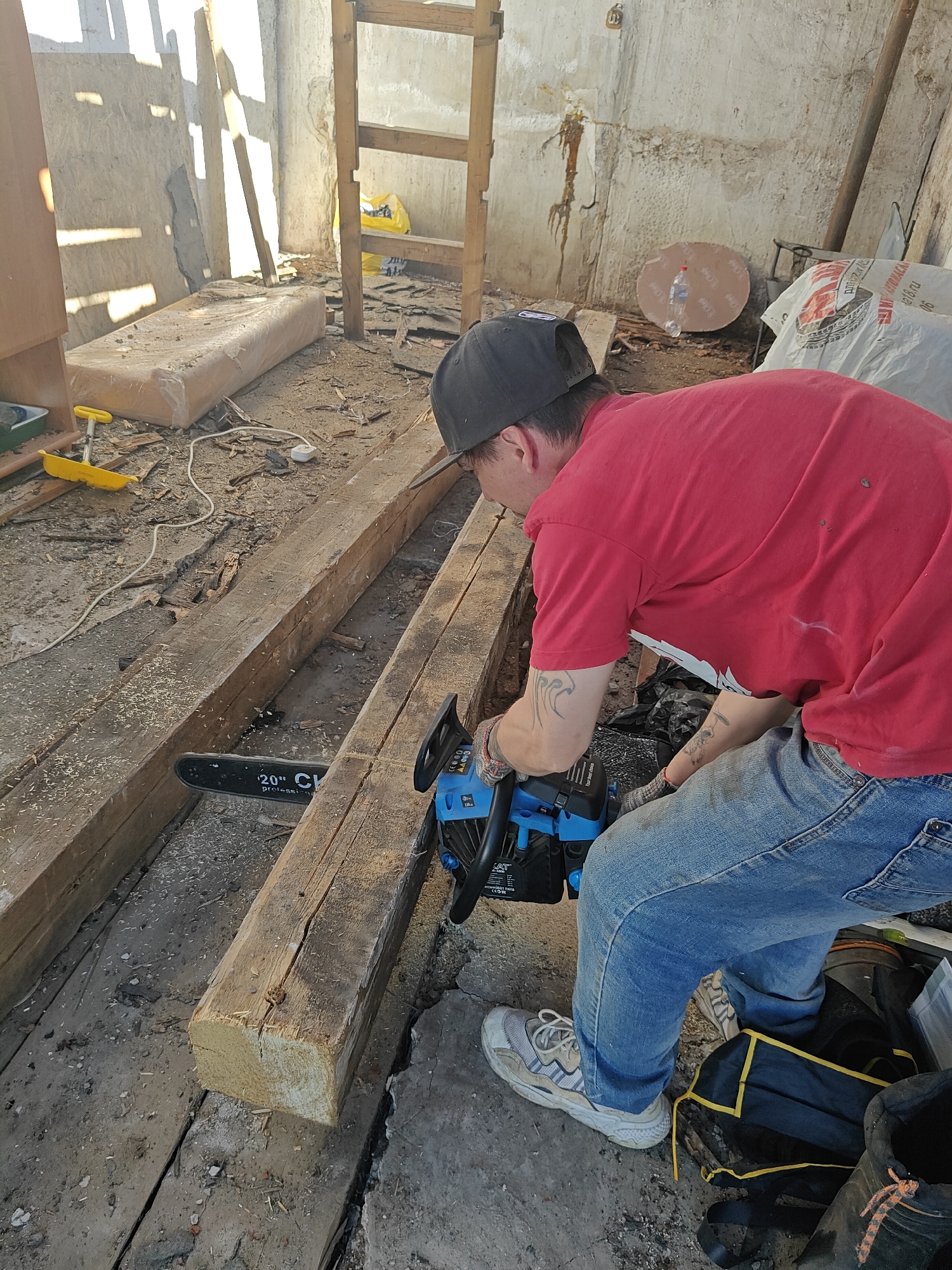 A man is using a chainsaw to cut a large wooden beam indoors. The man is wearing a red t-shirt, blue jeans, a black baseball cap, and white athletic shoes. He has tattoos on his arms. The chainsaw is blue and black. The wooden beam is lying on the floor, which appears to be made of weathered wood planks with debris scattered around. The room is cluttered with various items including a mattress, a yellow toy, bags, tools, and building materials. In the background, there is a wooden ladder and a wall with peeling paint and exposed concrete. The location is unspecified, but the image is described to be from the city of Chaikovsky, Russia. The man appears to be engaged in a construction or demolition activity.